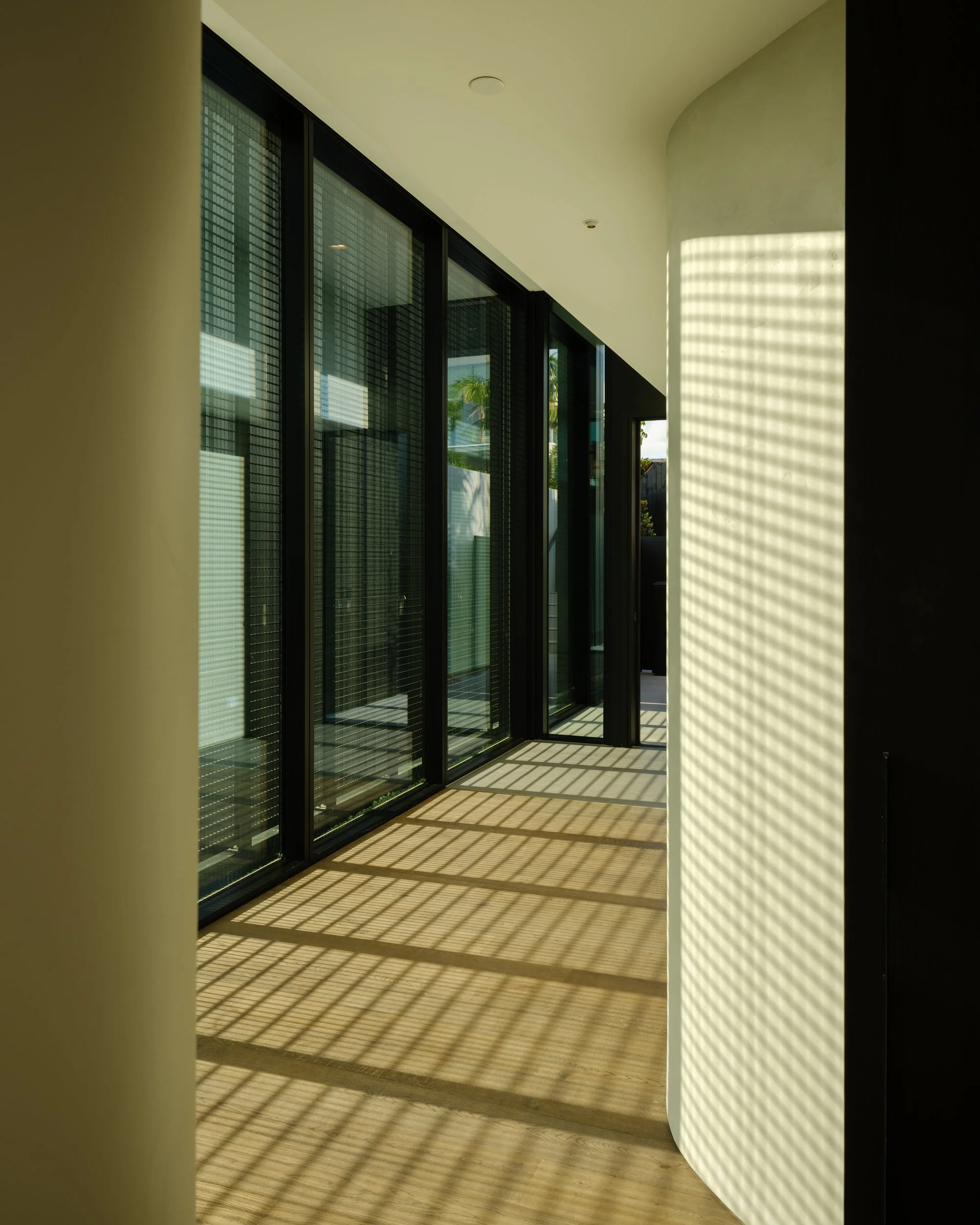 Sunlight casting striped shadows through a metal or wooden grid onto a wooden floor and a white curved wall in a modern interior space with glass doors.