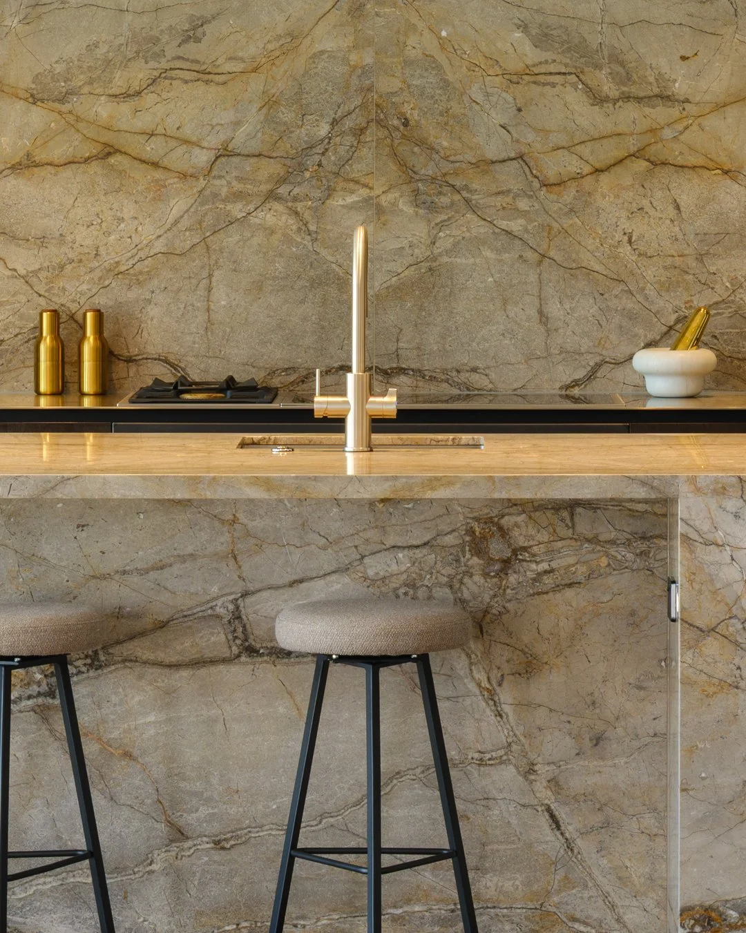 Modern luxury apartment kitchen with beige marble countertops and backsplash, a gold faucet, two gold bottles, a white bowl with a gold object, and two stools with beige cushioned seats and black metal legs.