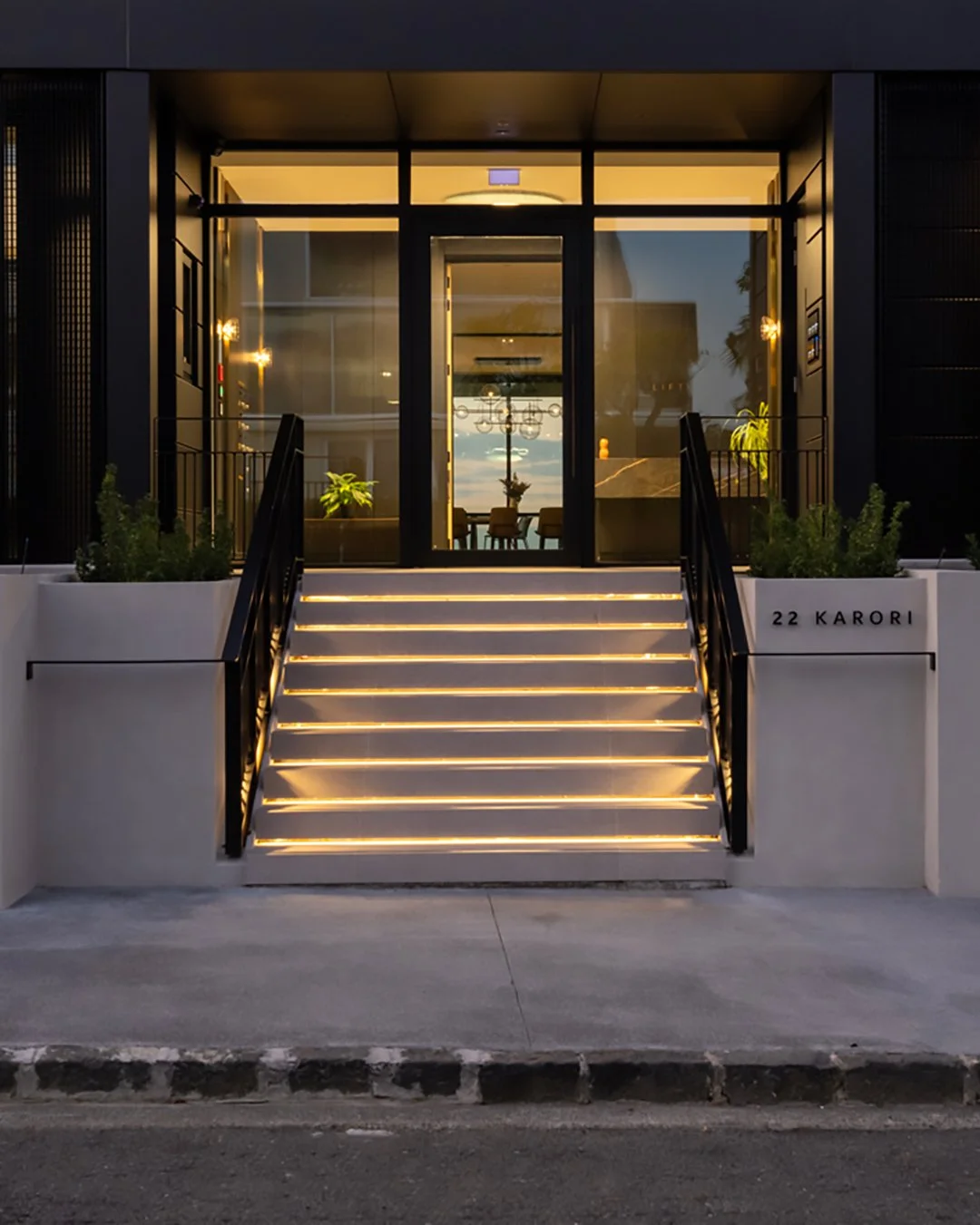 Modern luxury apartment building entrance with illuminated stairs, black railings, potted plants, and a glass front door showing a dining area inside. Address on wall reads '22 KARORI'.