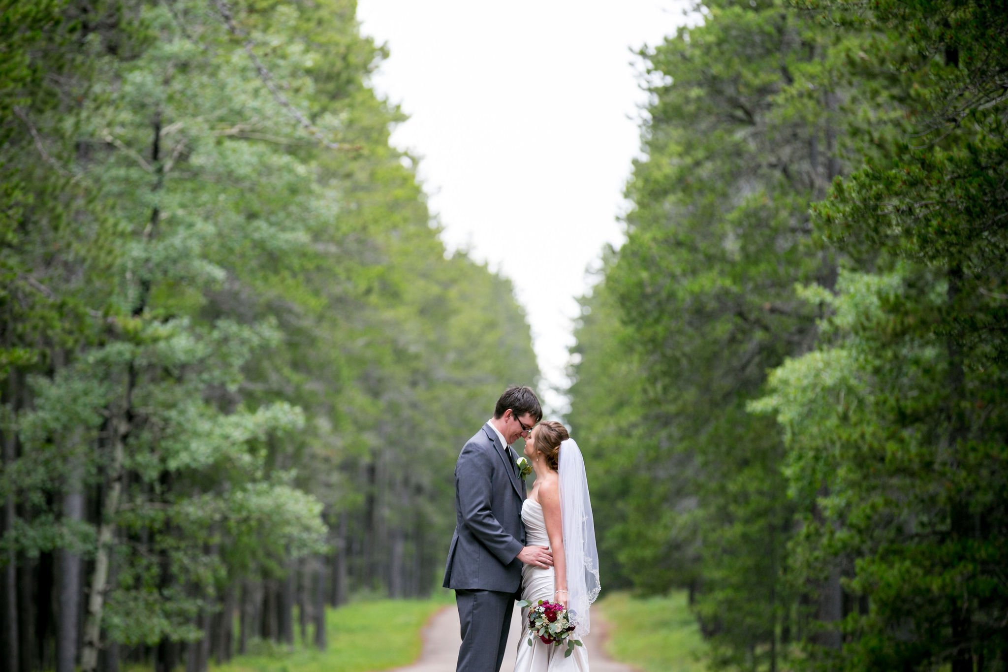Bride and groom embracing on a forest path, surrounded by trees.