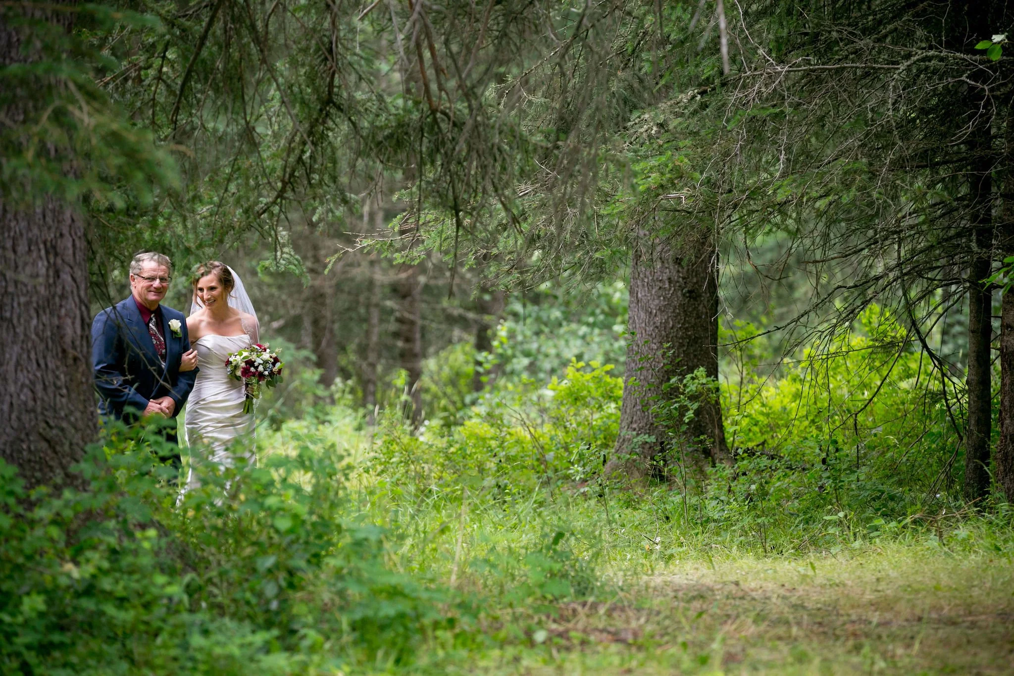 Bride and older man walking through forest, holding bouquet.
