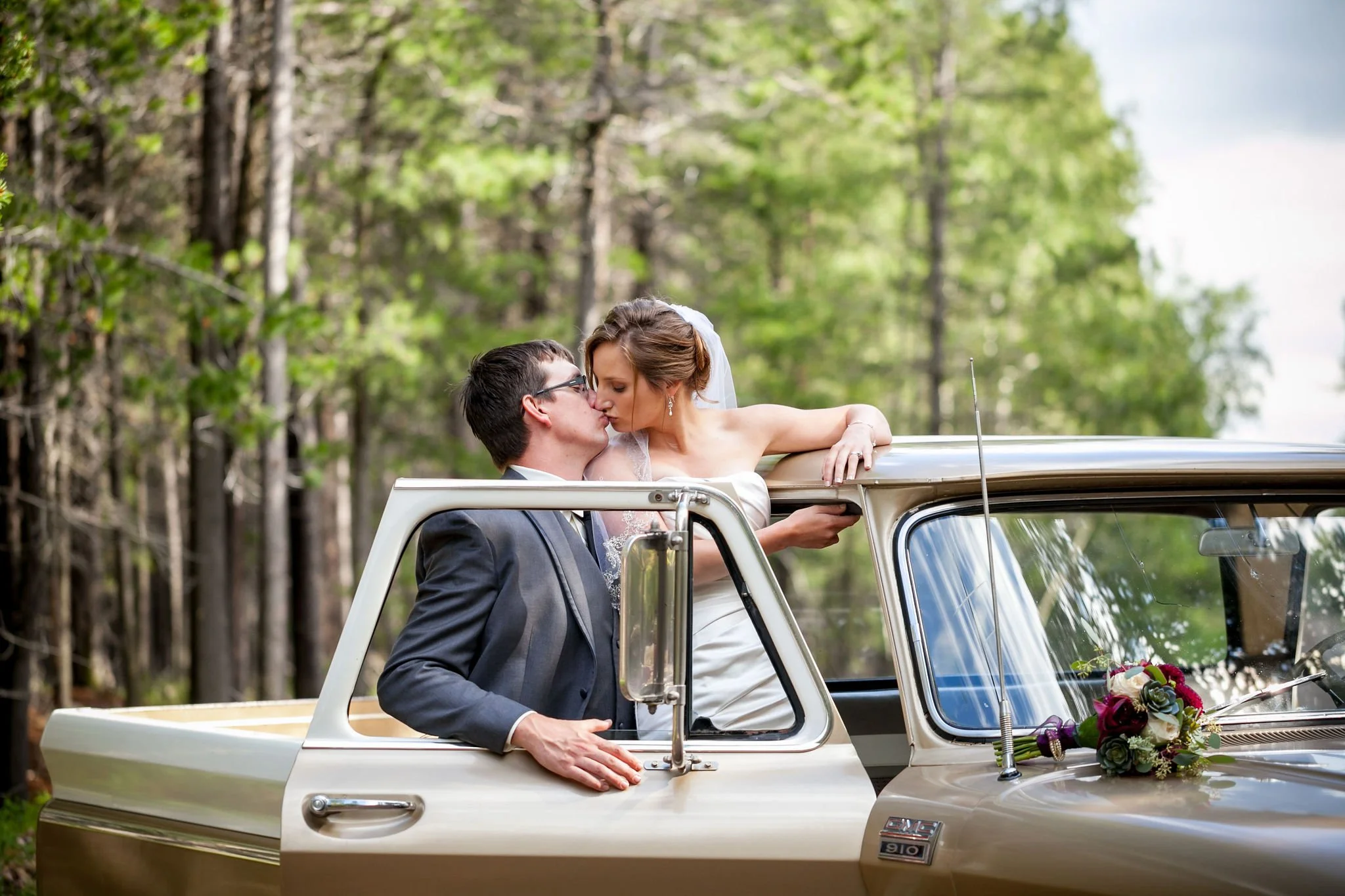 Bride and groom kissing in an open vintage pickup truck, with a bouquet on the hood, against a forest backdrop.