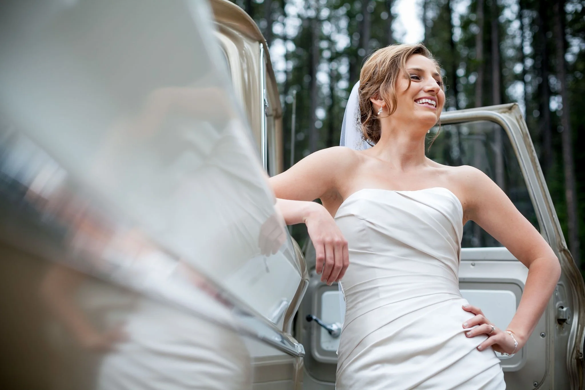 Bride in white dress smiling near vintage car in forest.