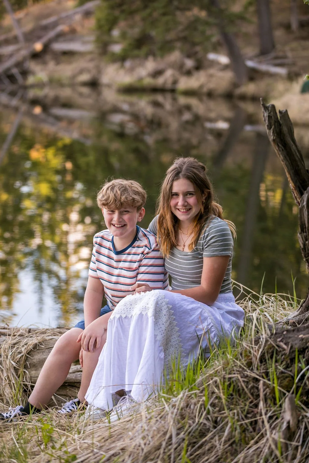 Two children sitting beside Loch Lomond, smiling, surrounded by grass and trees.