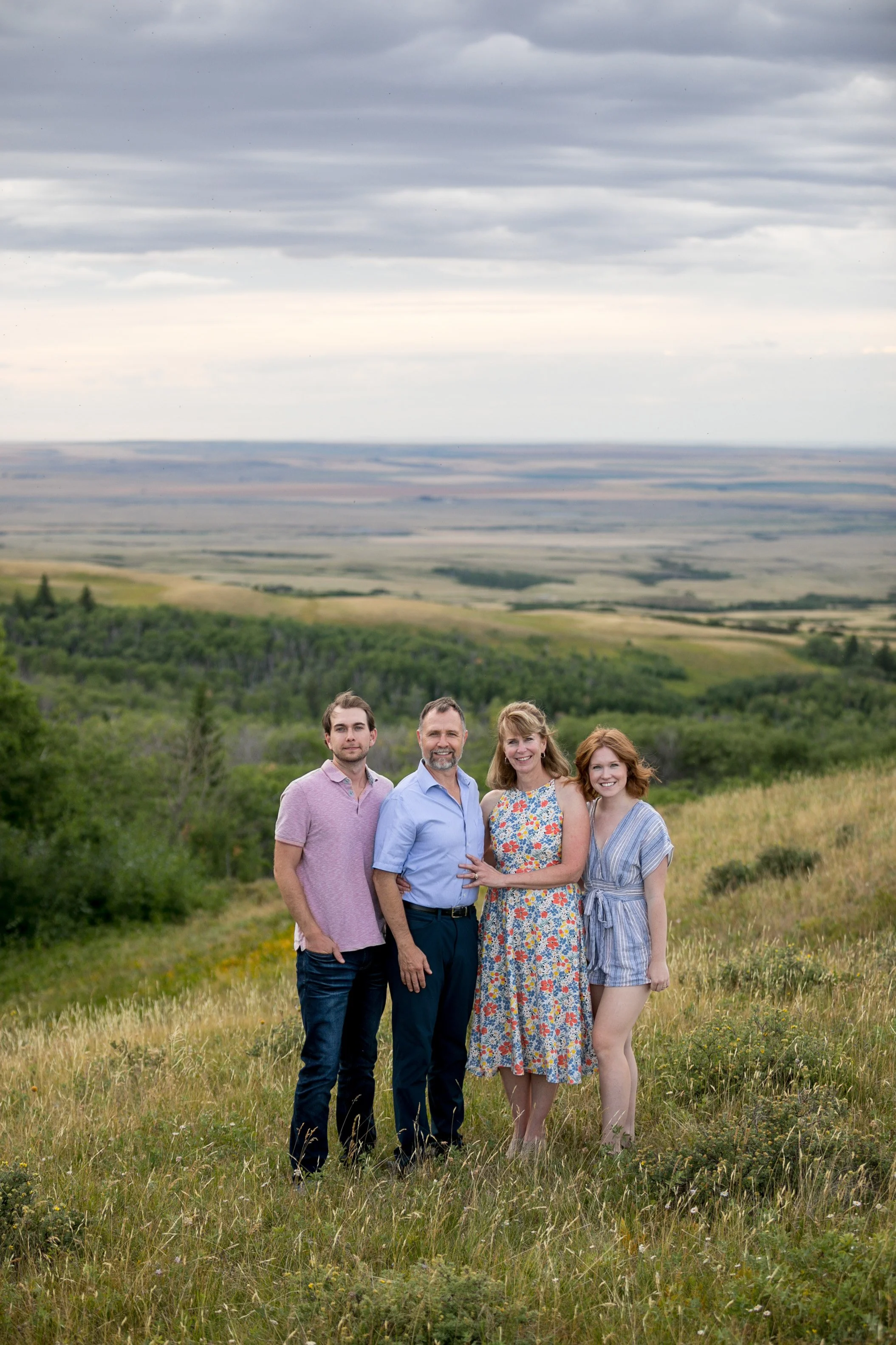 A family of four posing together on a grassy hillside with a scenic view of fields and a cloudy sky in the background.