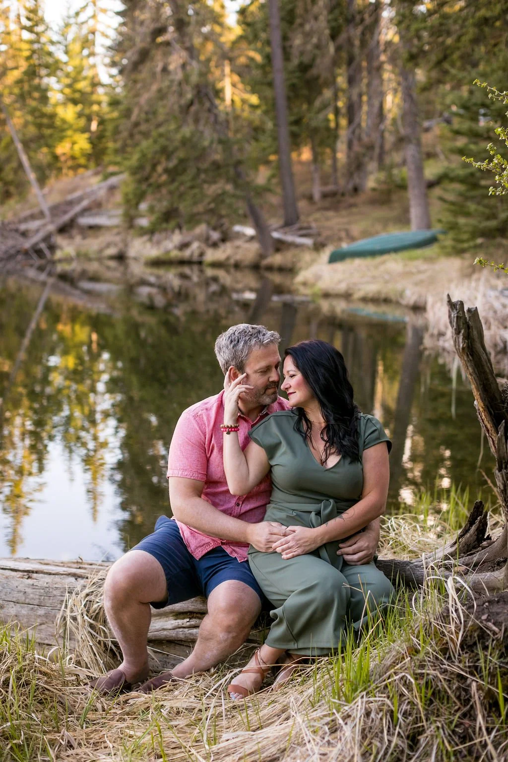 A couple sitting on a log beside Loch Leven in Cypress Hills Interprovincial Park, embracing and gazing at each other affectionately. The background features trees and a calm water surface.