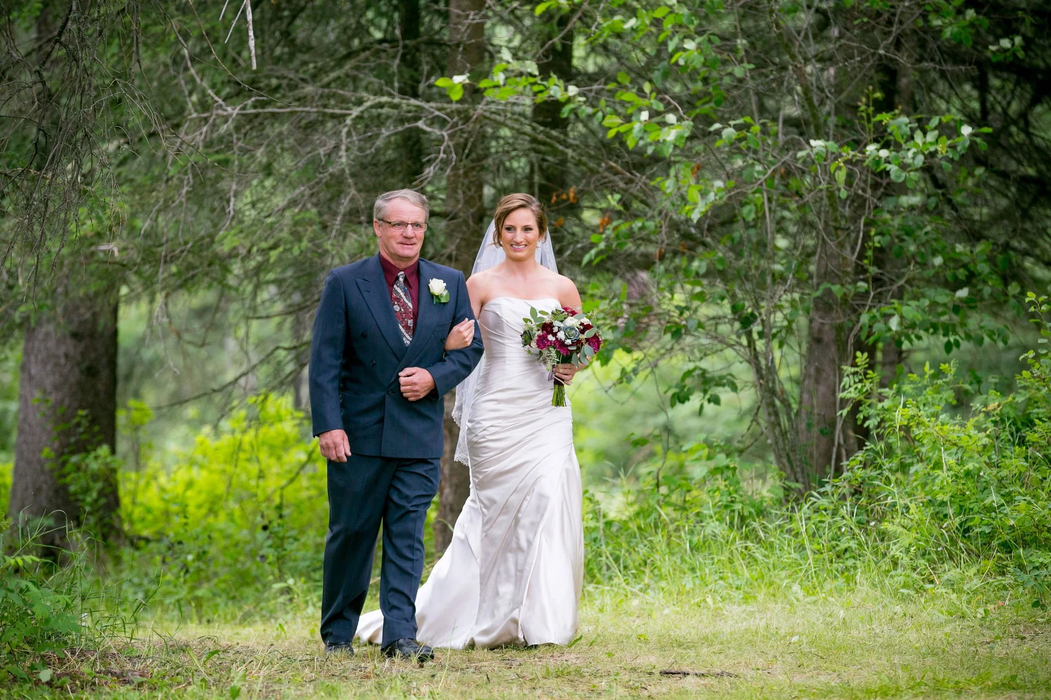 Bride walking with an older man outdoors, surrounded by trees, both dressed formally.