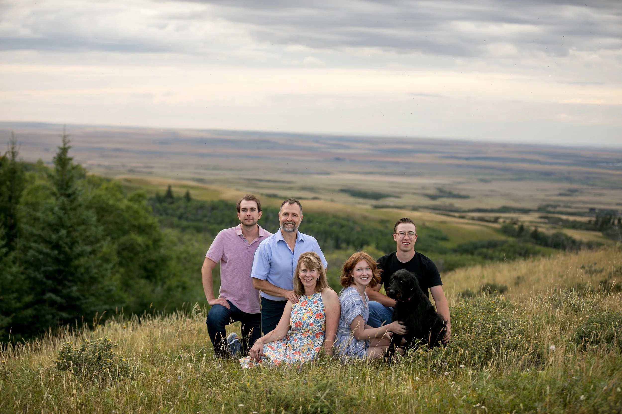 Family and dog sitting in a grassy field with rolling hills in the background.
