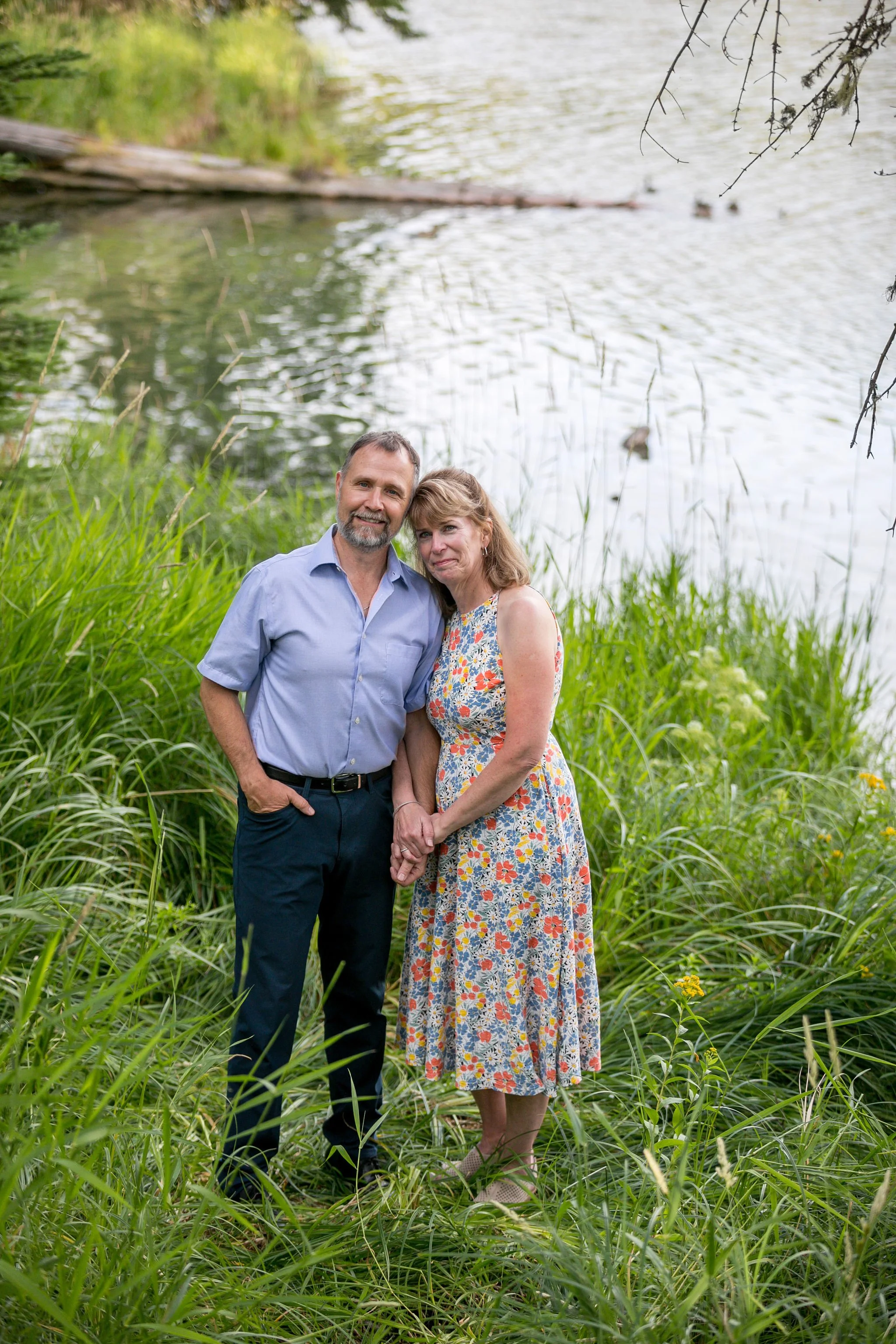 A couple standing by a lakeside, surrounded by green grass, holding hands and smiling. The man is wearing a light blue shirt and dark pants, and the woman is in a floral dress. Trees and water are in the background.