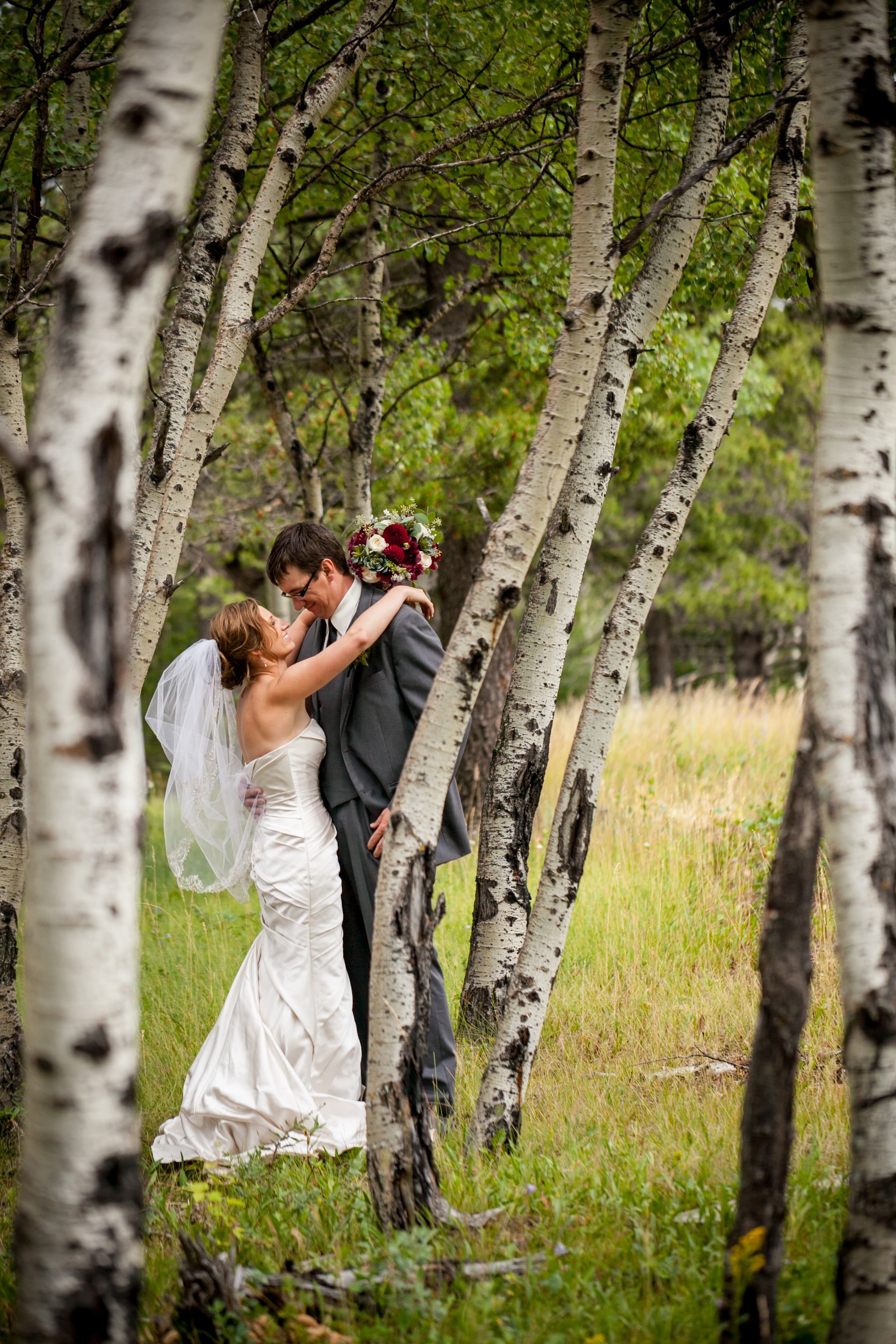 Bride and groom embracing in a forest with birch trees and green foliage, bride holding a bouquet.