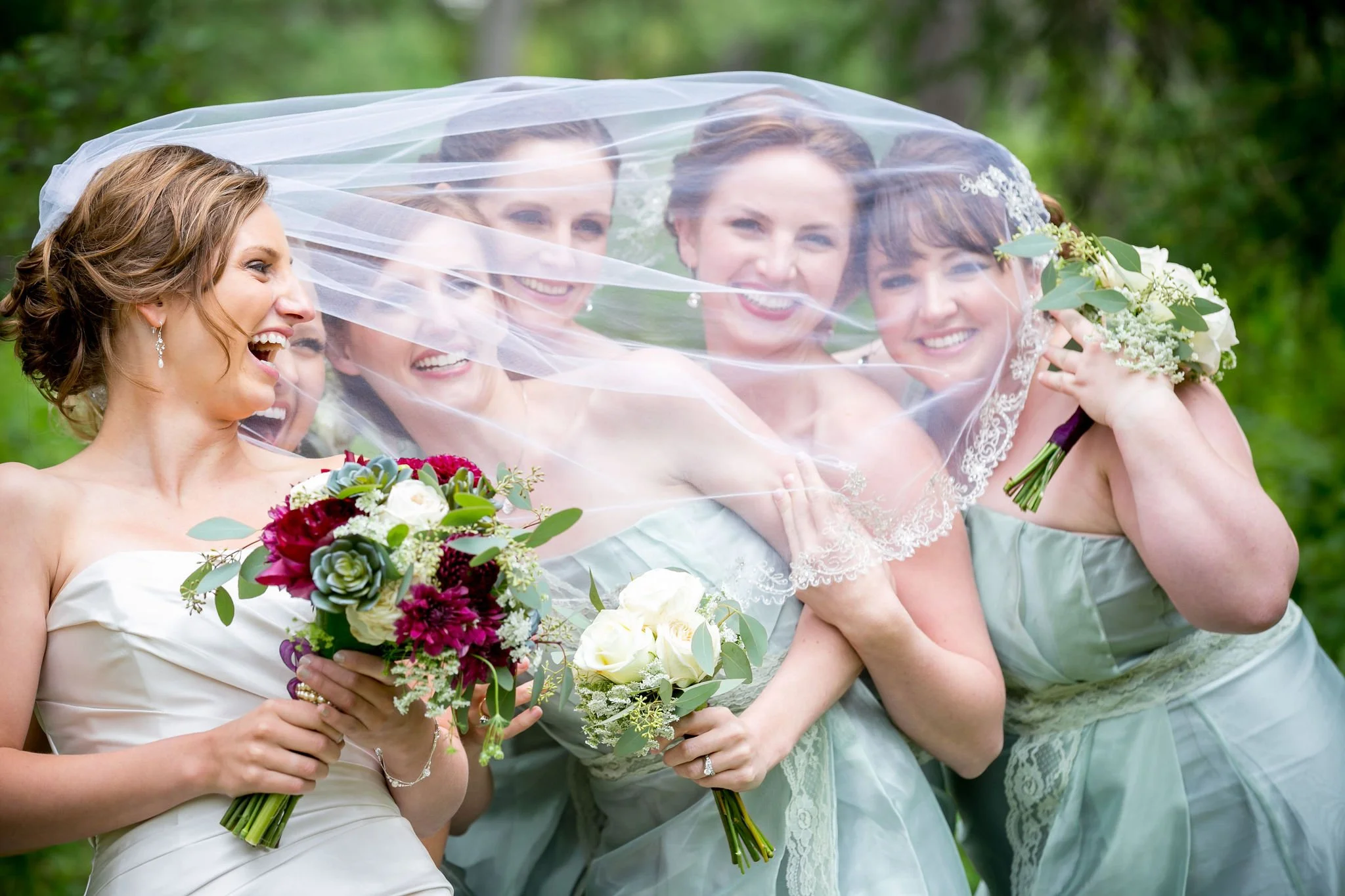 Bride and bridesmaids laughing under veil, holding flowers.