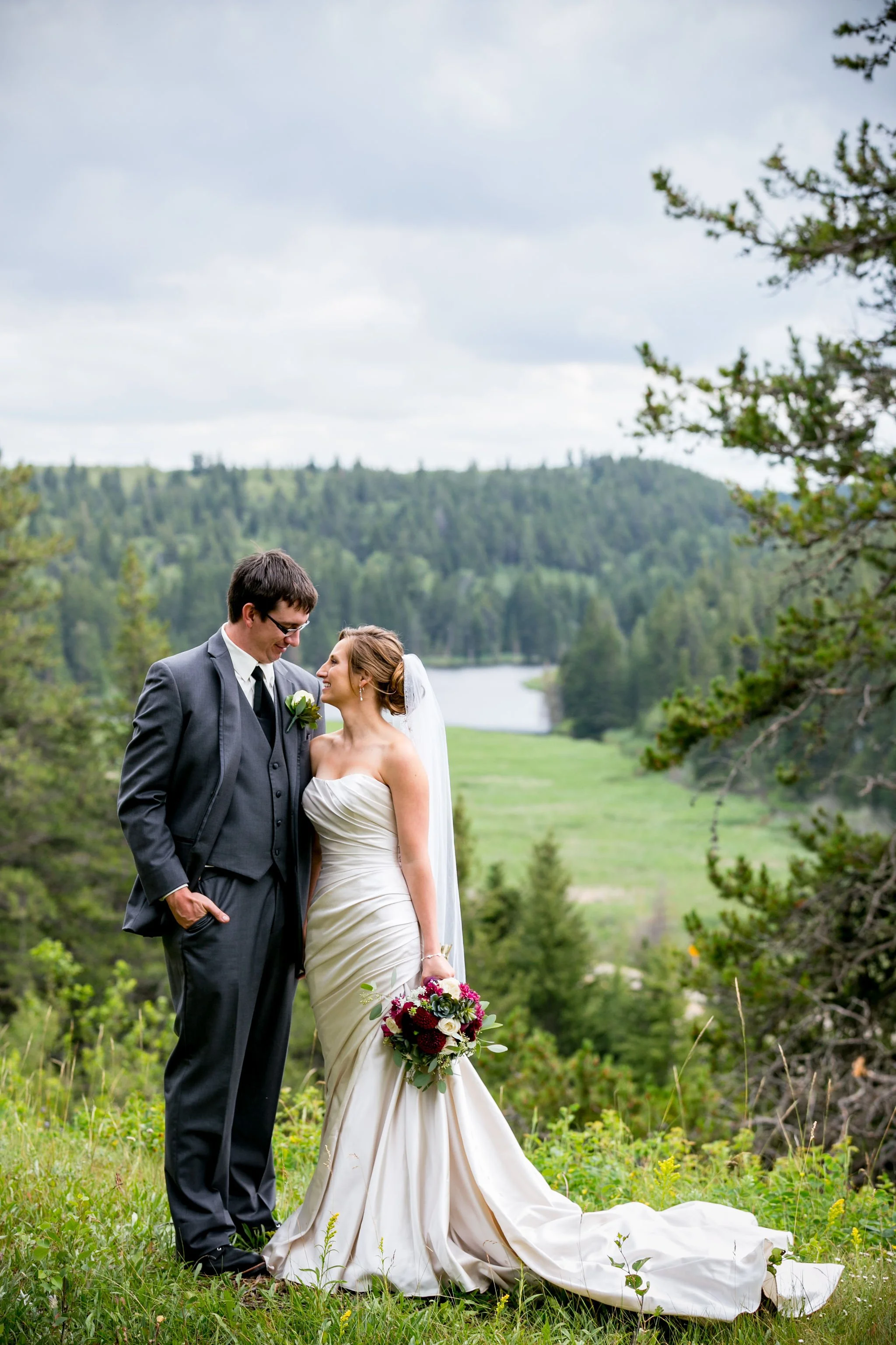 Bride and groom standing on grassy landscape with trees and a lake in the background.