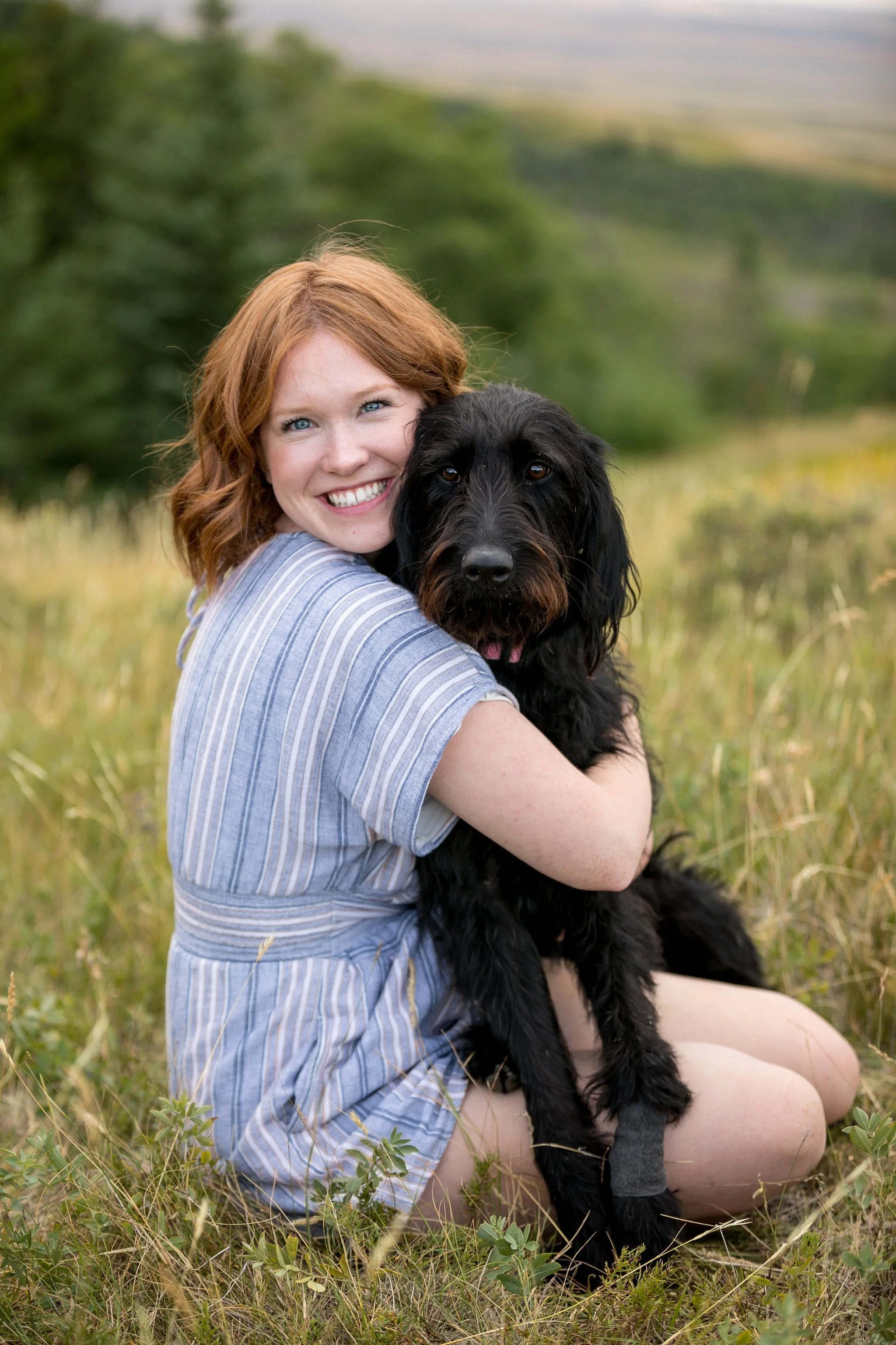 Woman in blue-striped dress hugging a black dog in a grassy field with trees in the background.
