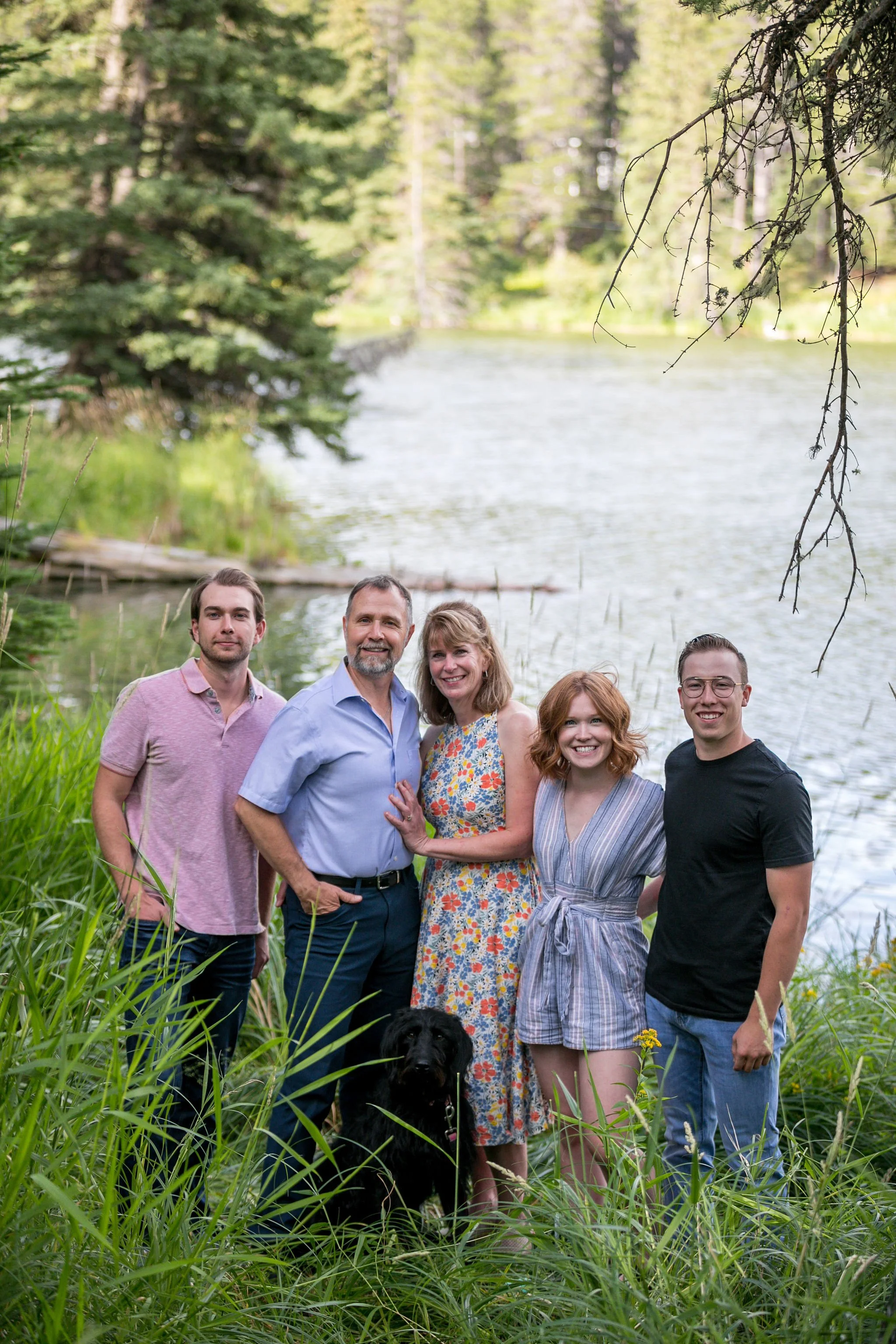 Family posing by a lake with a black dog, surrounded by trees and greenery.