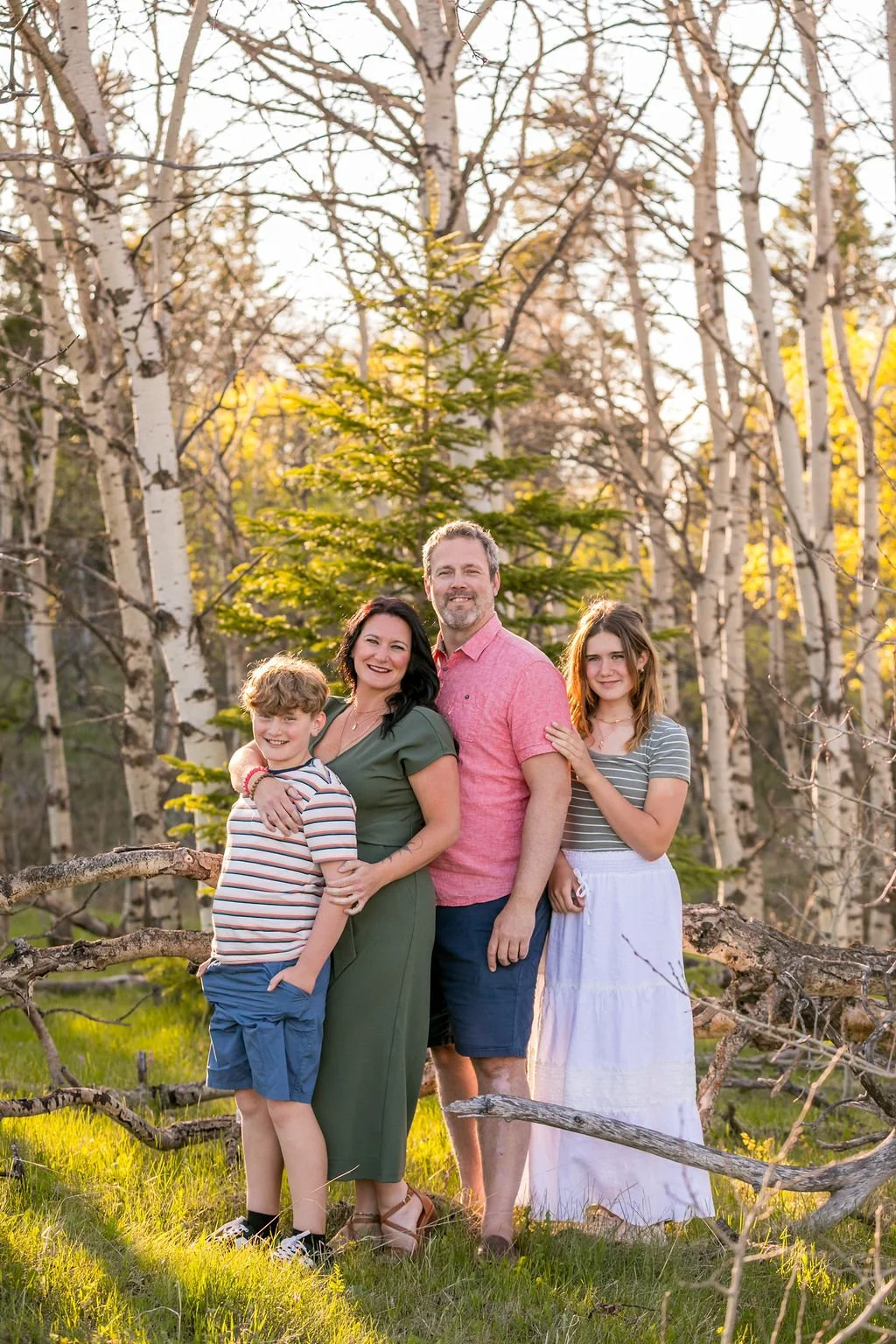 A family of four posing outdoors for their family photo in Cypress Hills Provincial Park Saskatchewan. The group includes a mother, father, and two children, standing close together and smiling. The scene is lit by natural sunlight.
