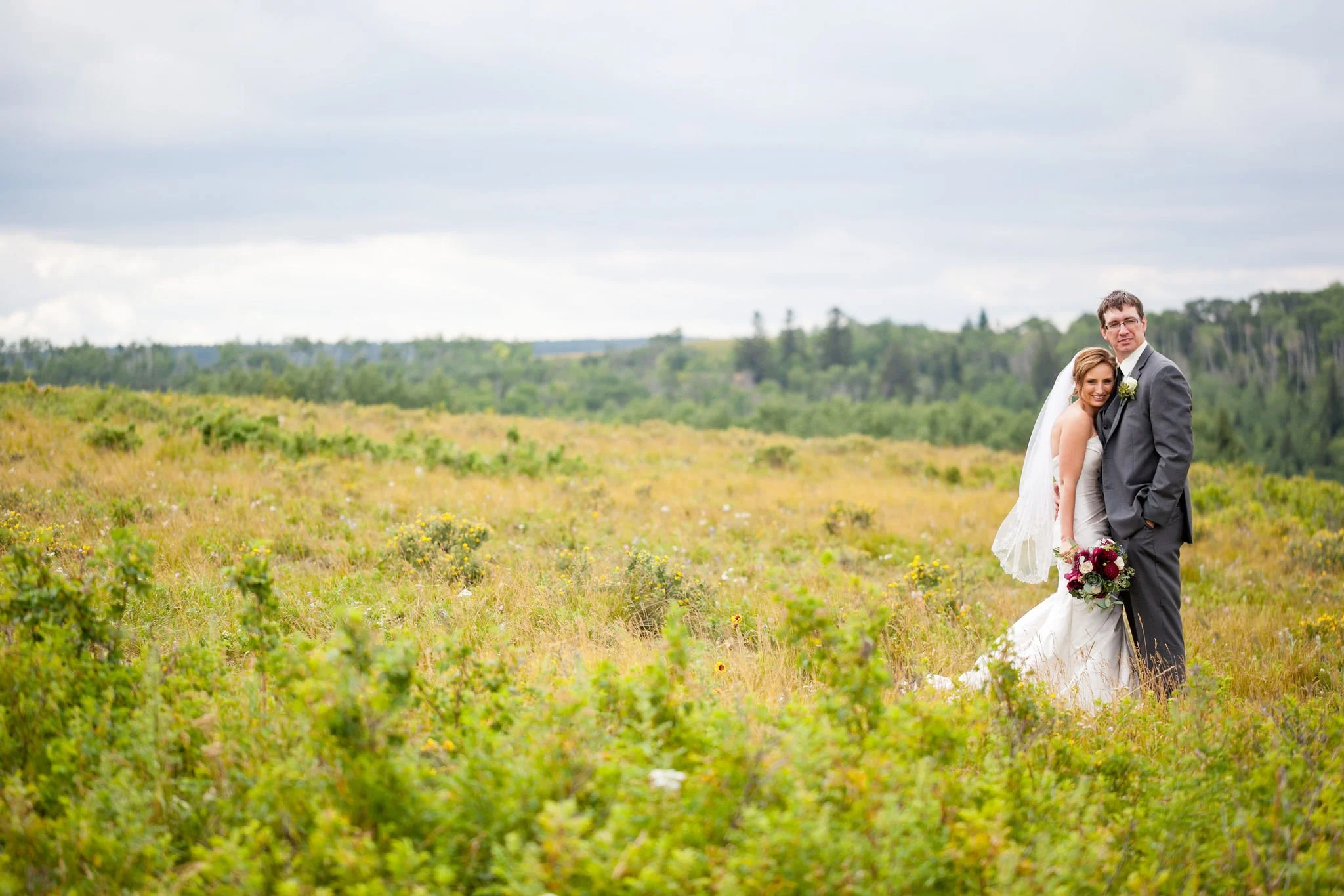 Bride and groom posing in a grassy field with a forested background.
