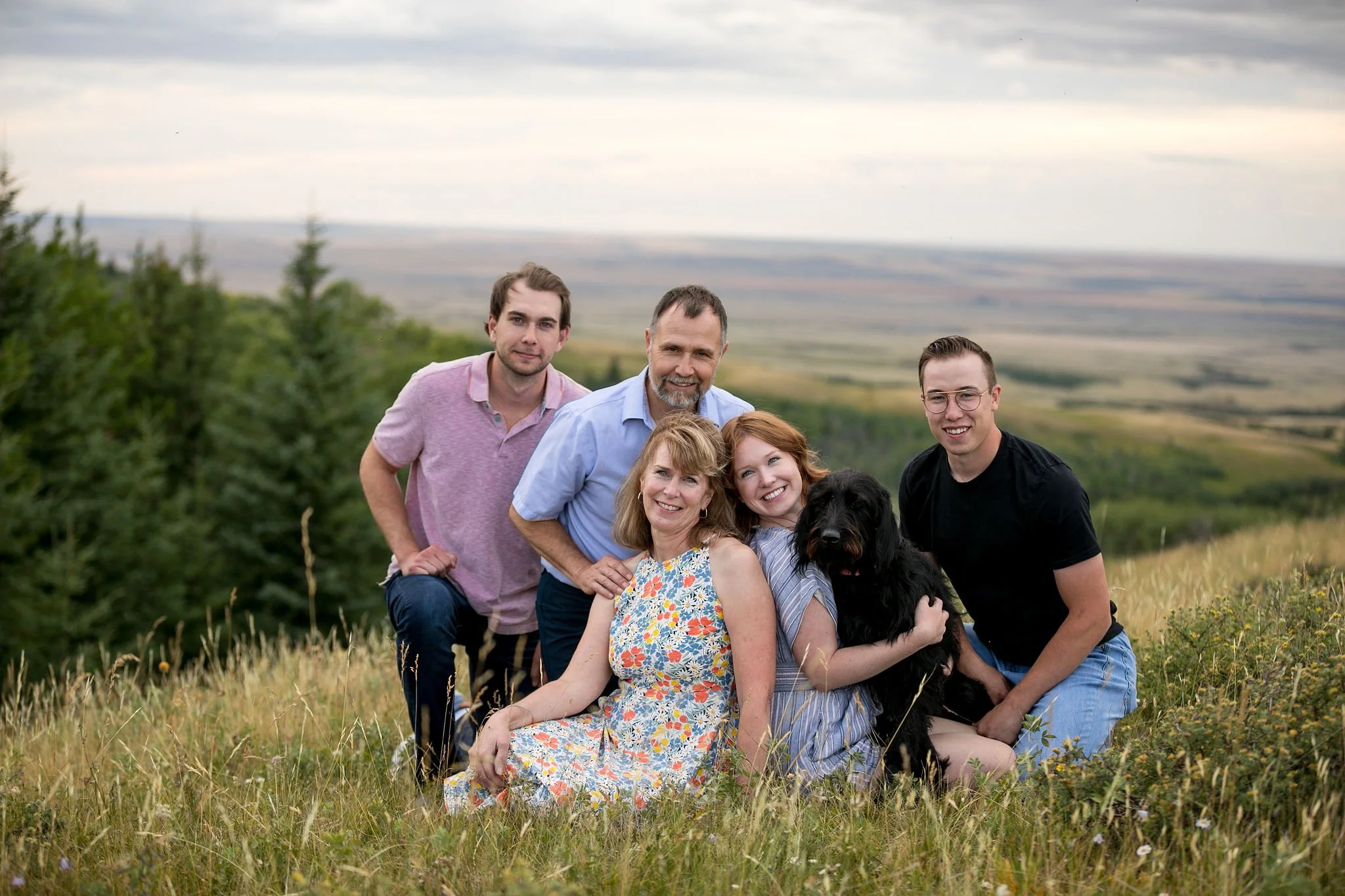 Family and dog sitting in a grassy field with a scenic background.