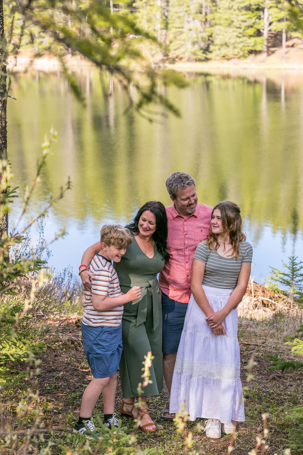 A family of four standing in a forest setting. The group includes two adults and two children, smiling and interacting with each other. The lake reflects the surrounding pine trees, and the scene conveys a sense of a nature outing or family picnic.