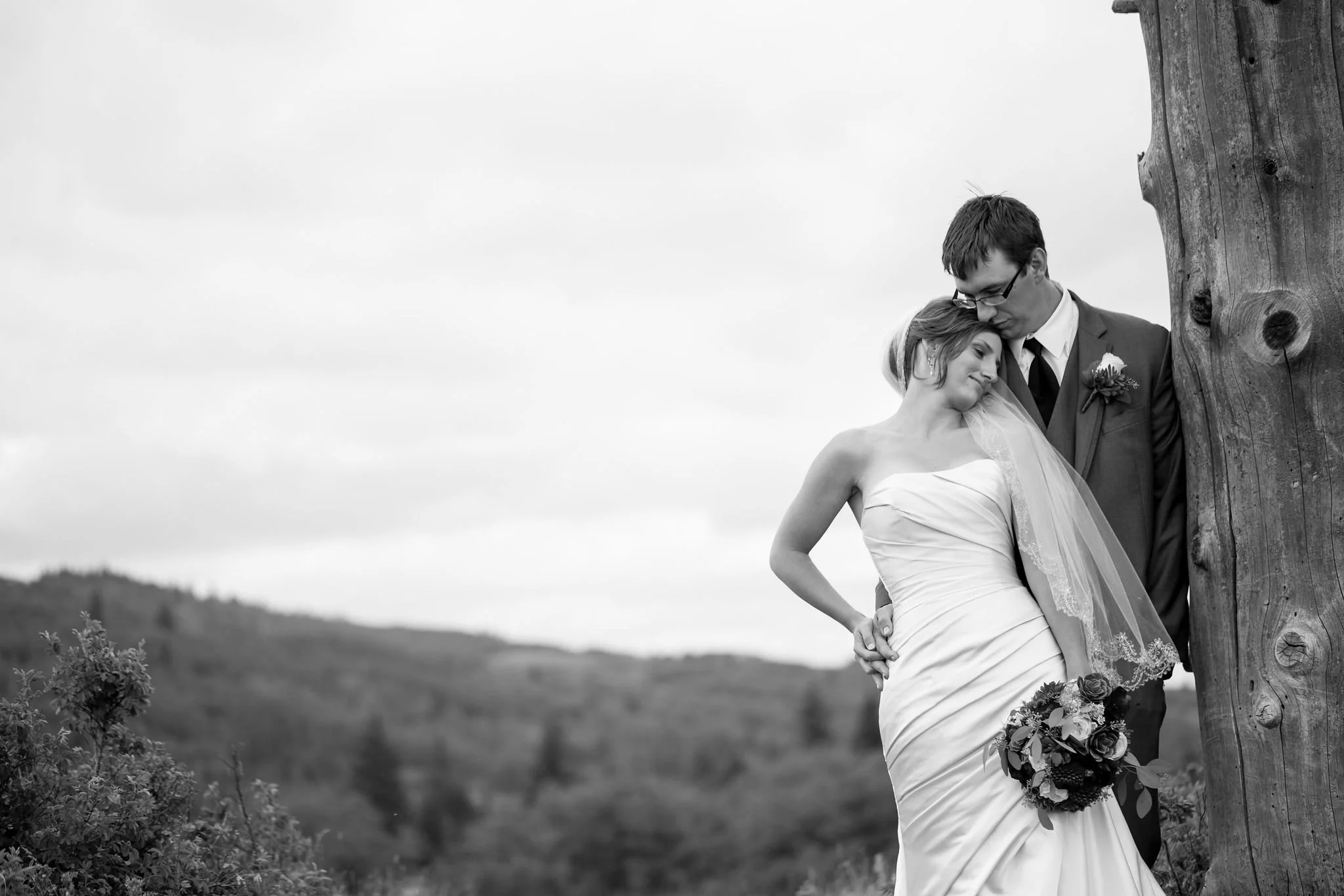 Black and white photo of a bride in a strapless gown holding a bouquet, leaning against a groom in a suit, standing beside a tree, with a scenic landscape backdrop.