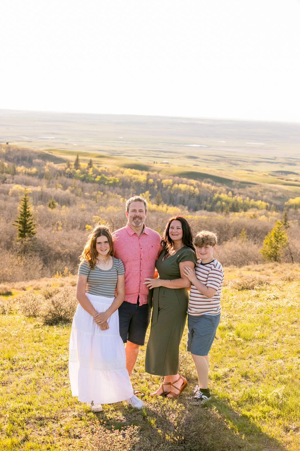 Family posing for their family photograph standing in a grassy field with trees  overlook lookout point at Cypress Hills Interprovincial Park. 