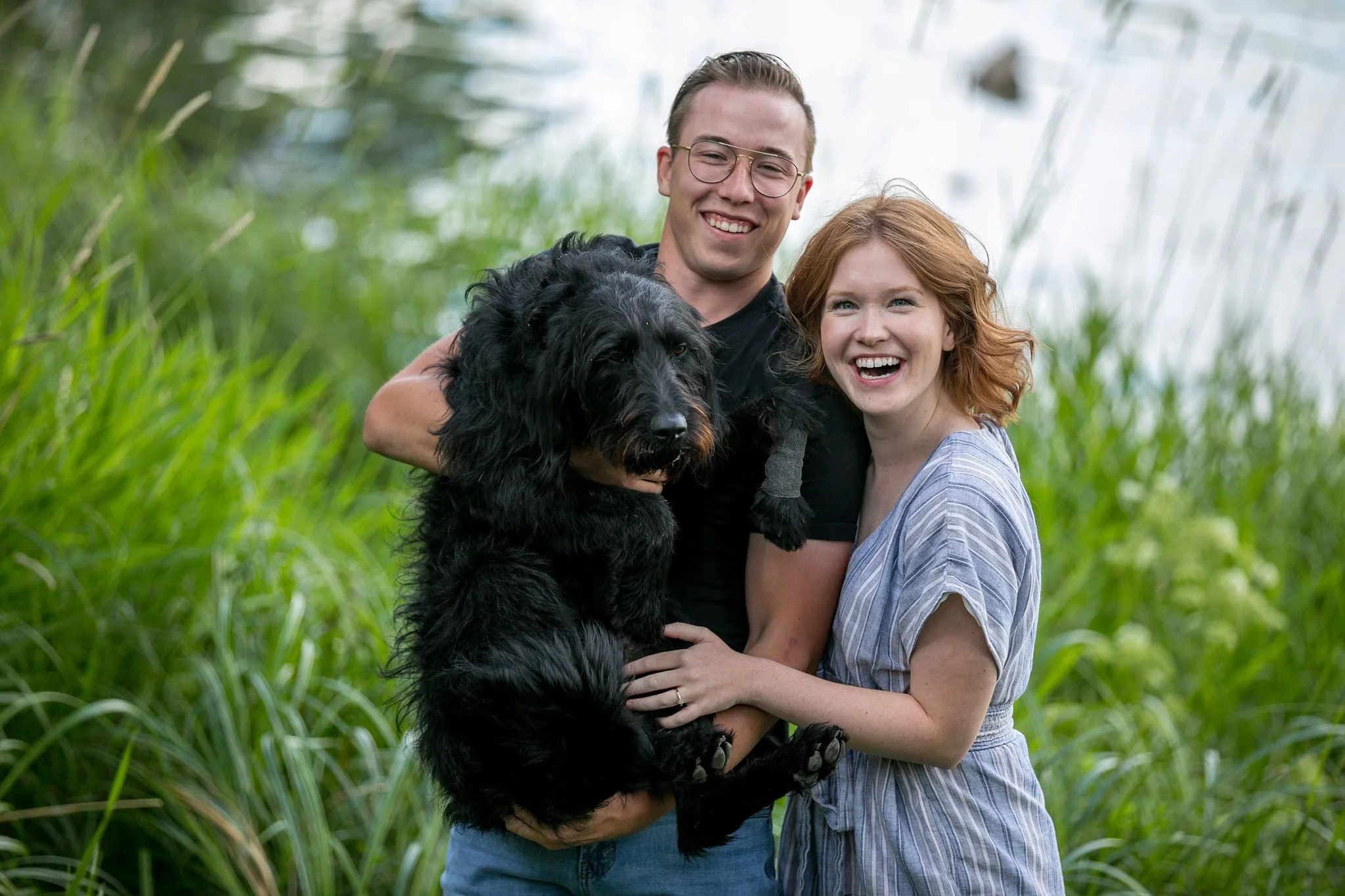A smiling couple holding a black dog in a grassy park.