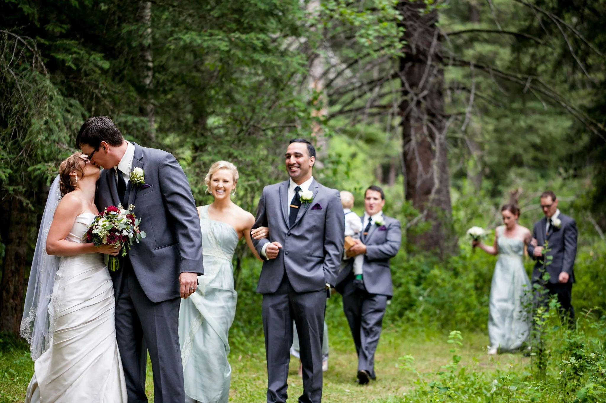 Wedding party walking in a forest, bride and groom kissing, others following, wearing formal attire.