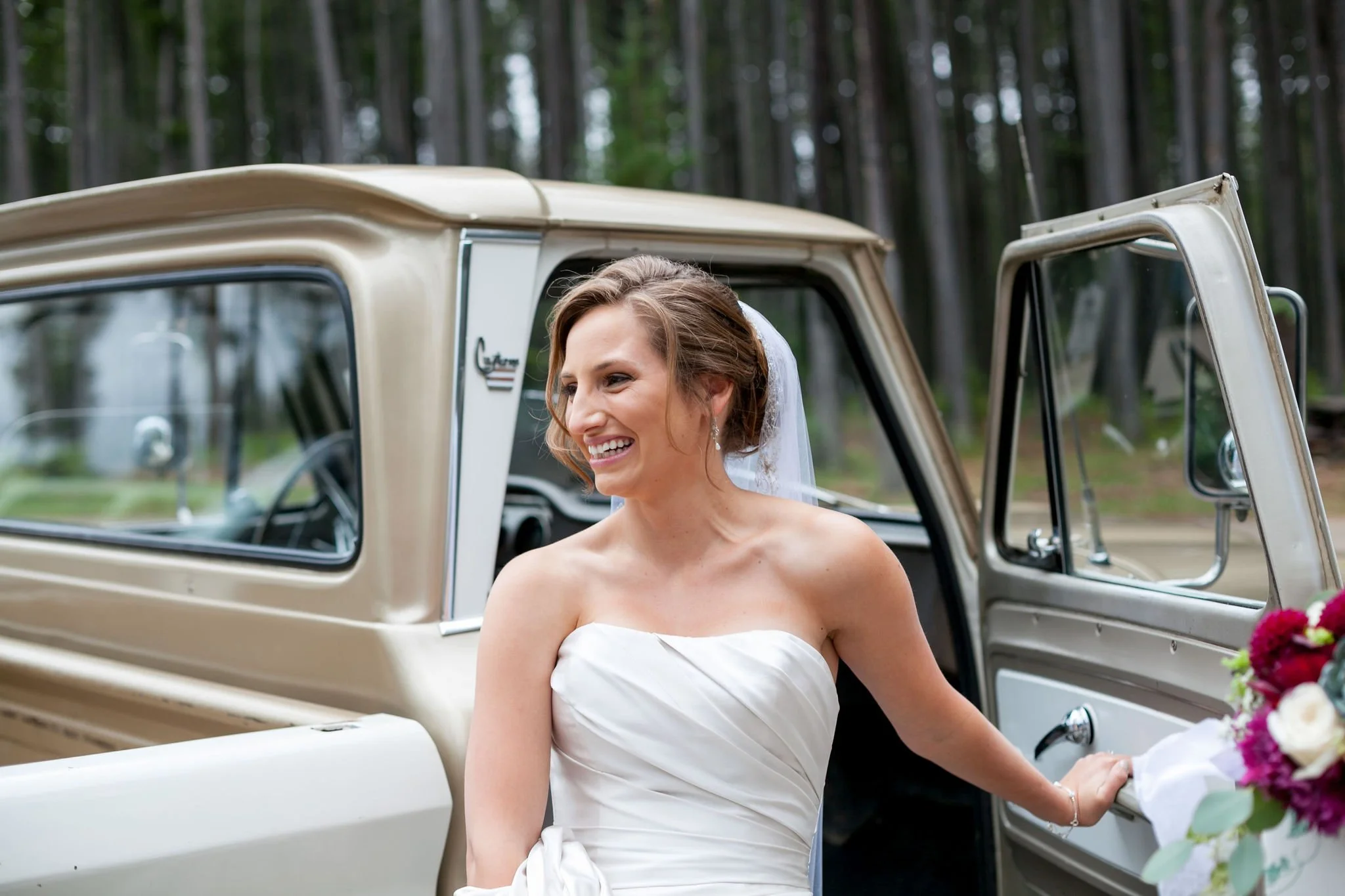 Bride in white dress smiling near vintage truck and flowers in wooded area.