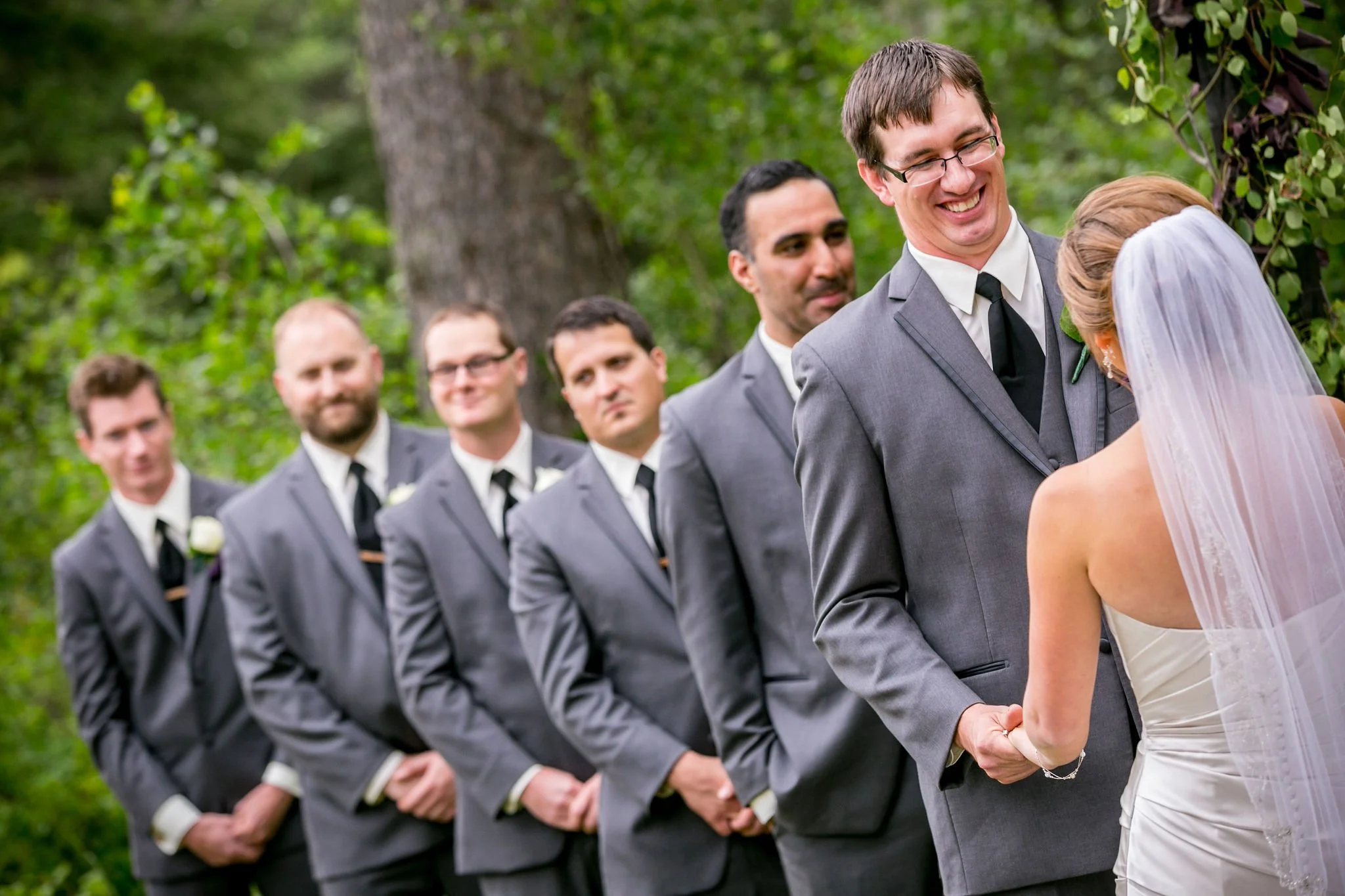 Bride and groom holding hands during outdoor wedding ceremony, with groomsmen in gray suits standing in line.