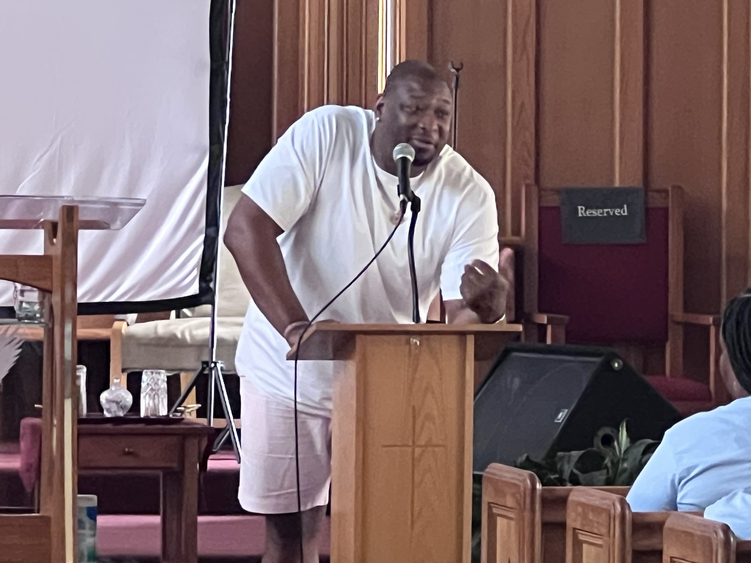 A man in a white shirt and shorts speaking passionately at a podium in a church or auditorium, gesturing with his right hand.