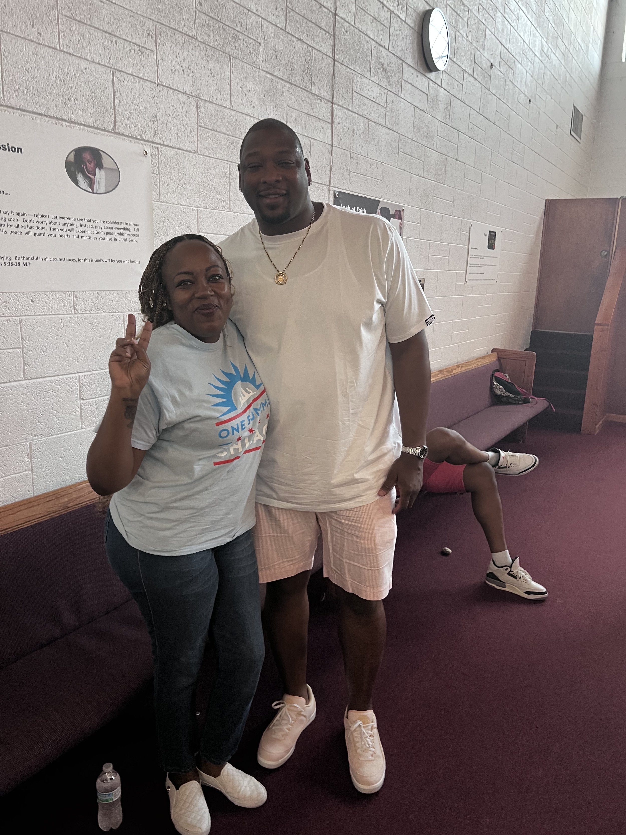 Two people standing next to each other inside a building, one woman and one man, both smiling and making a peace sign. A third person is seated on a bench in the background.