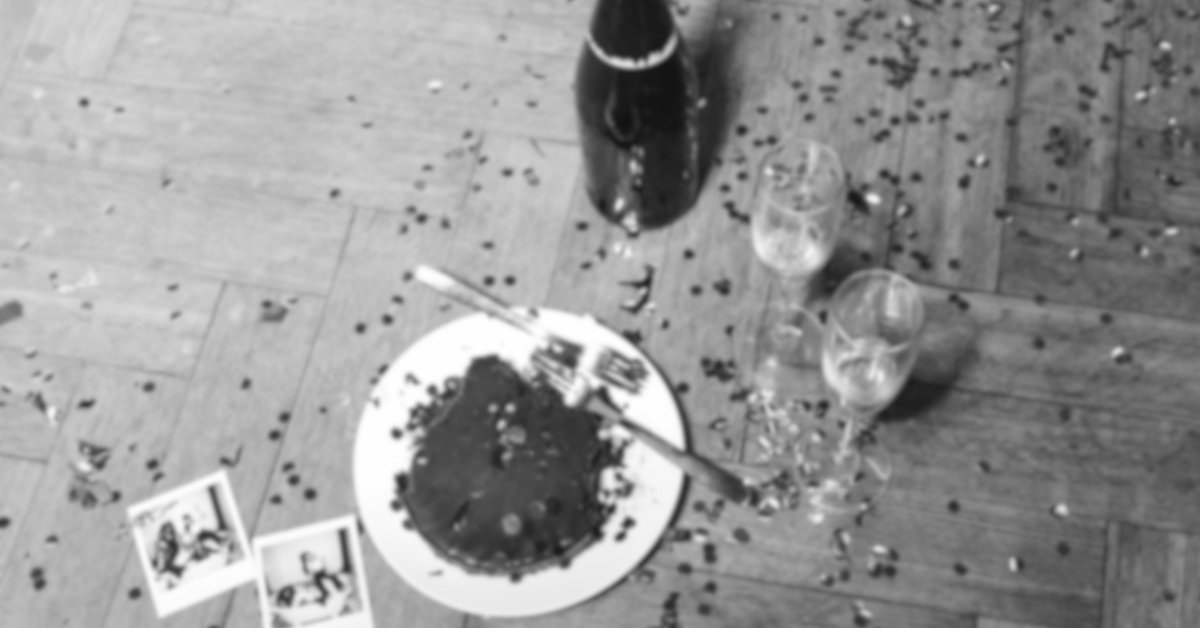 Empty party table with a plate of leftover cake, two empty wine glasses, a bottle, scattered confetti, and two Polaroid photos on a wooden floor.