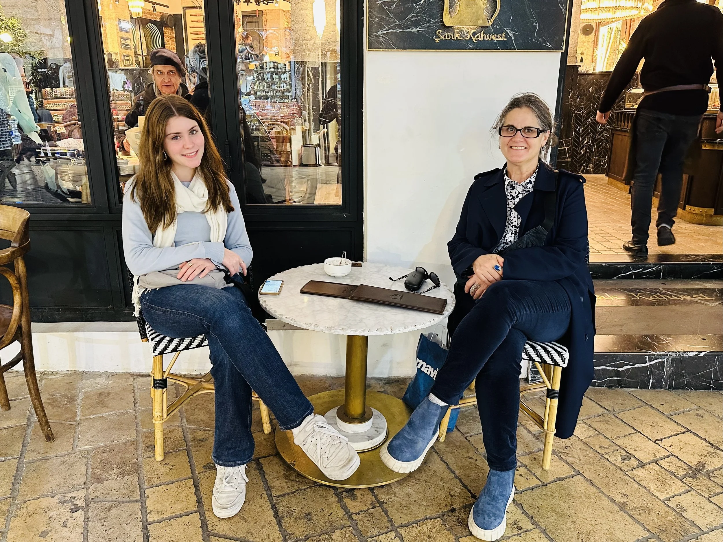 Two women sitting at a small round marble-top table inside a cafe. One woman has long brown hair, wearing a light blue sweater, white scarf, and jeans. The other woman has gray hair, glasses, and is wearing a navy blue coat and boots. On the table are a brown notebook, a pair of sunglasses, a black car key, and a small bowl. In the background, a large window reveals a woman in a black beret looking inside.
