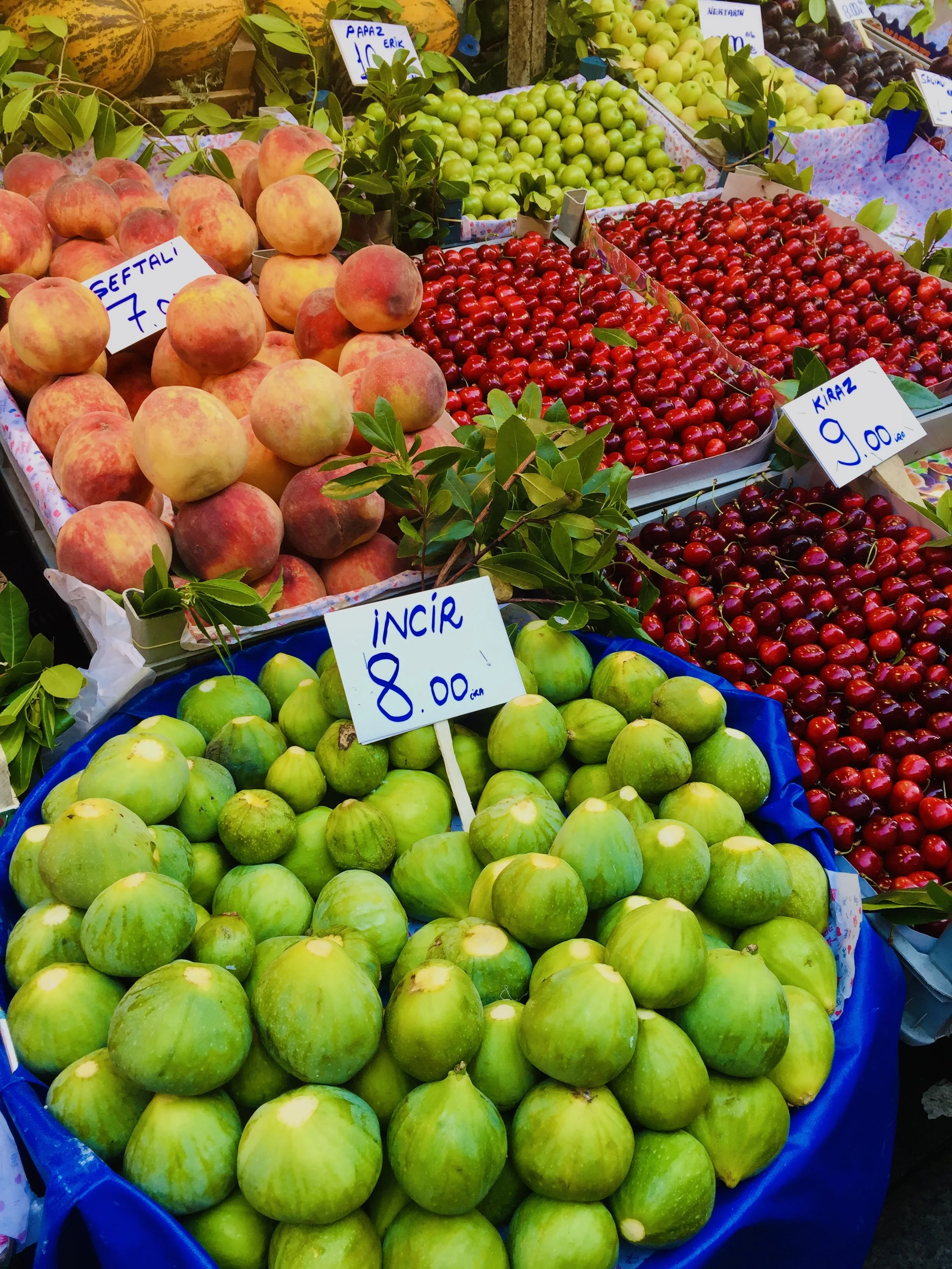 Fresh green figs, peaches, and cherries displayed in market stalls with handwritten price tags.