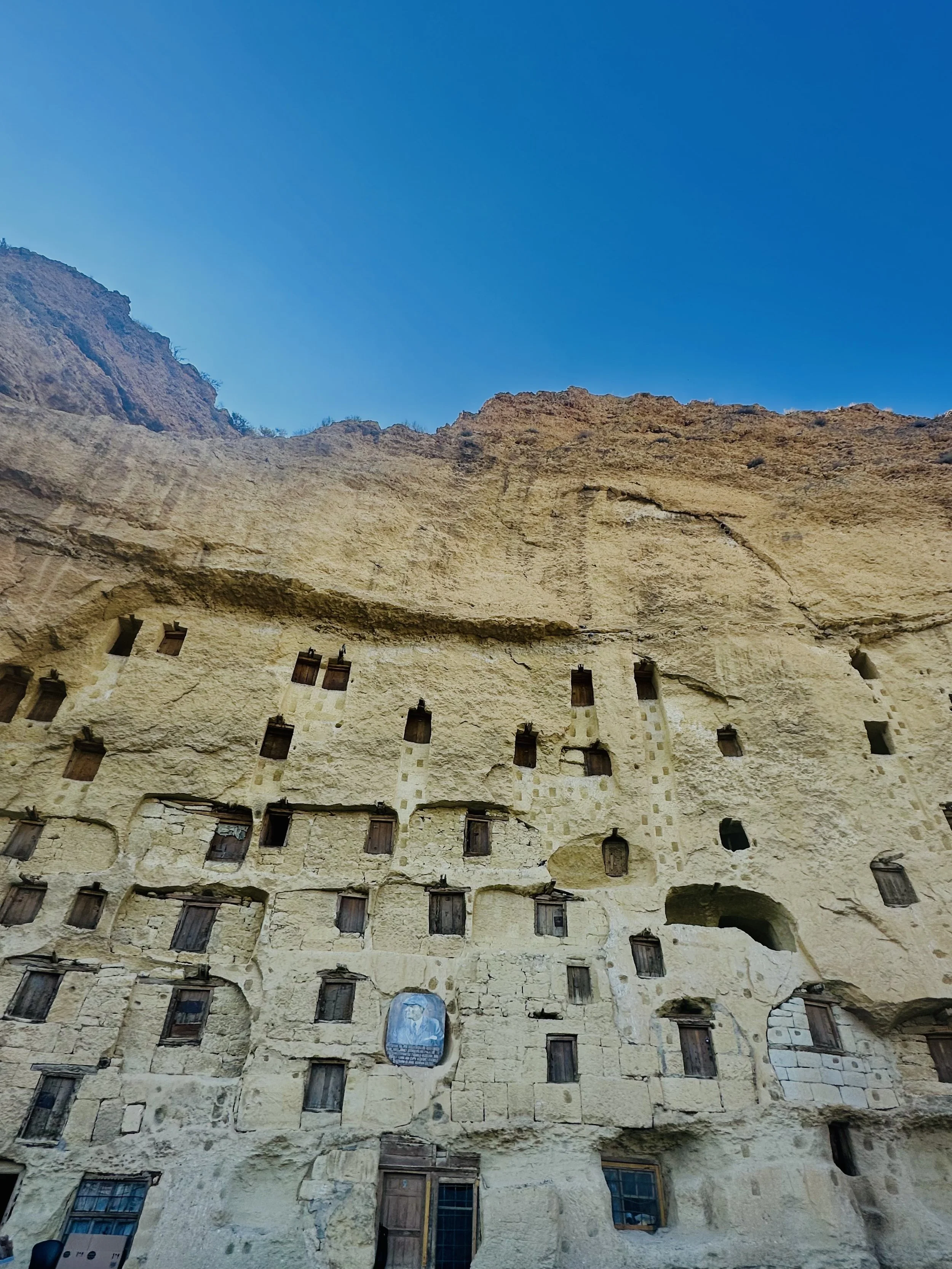 Ancient cliffside dwelling with small wooden windows built into a rocky face under a clear blue sky.
