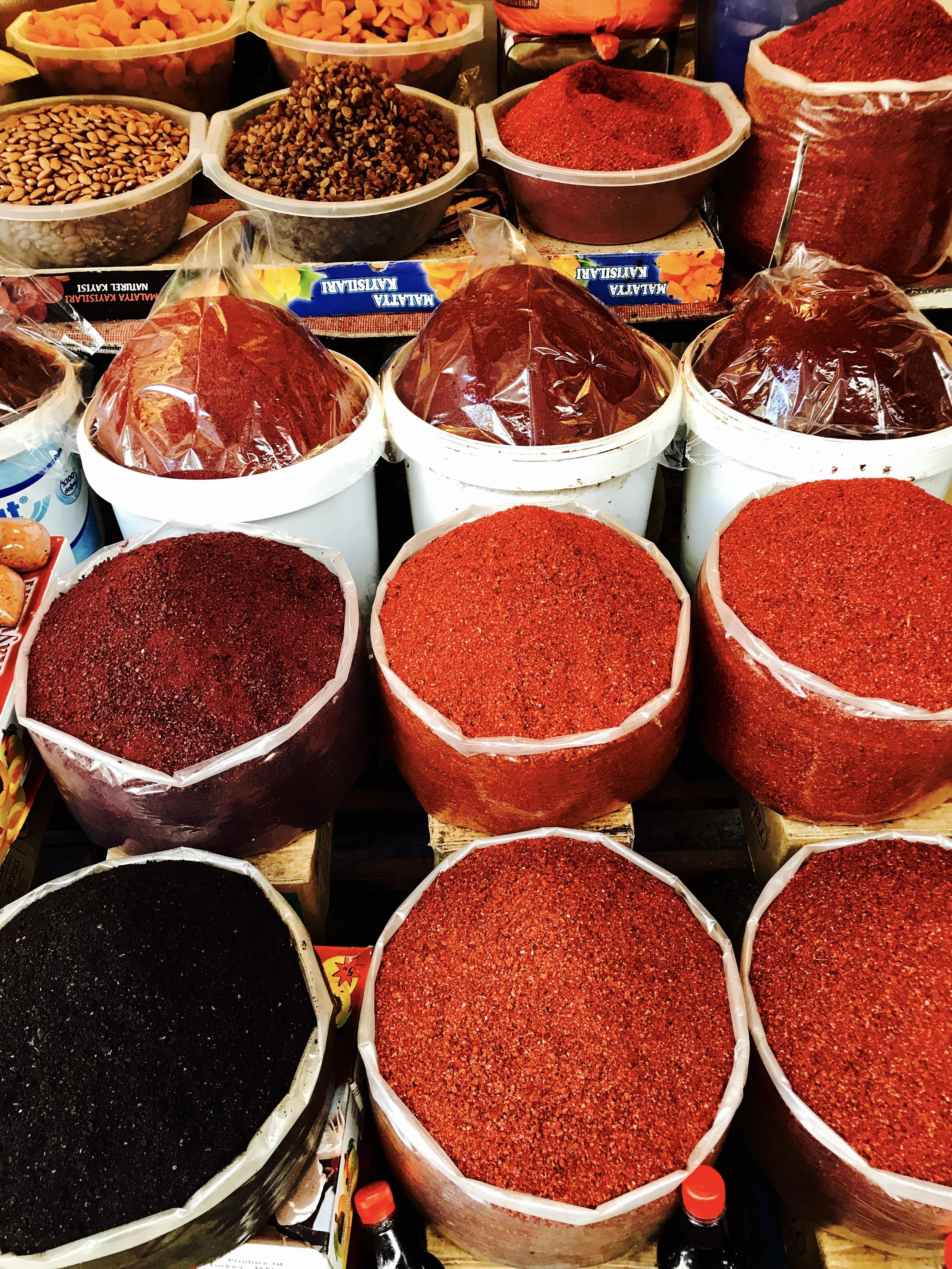 Various spices and herbs displayed in large open containers, including red, black, and brown ground spices.