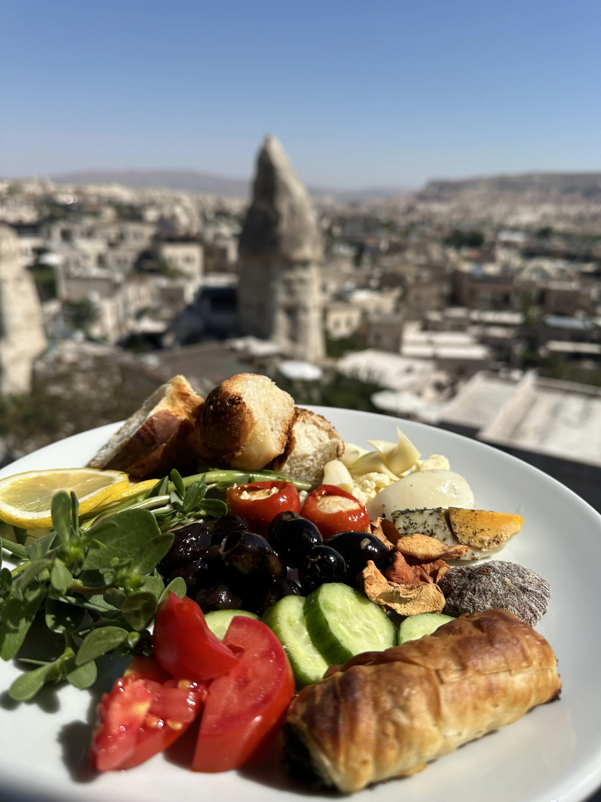 A plate of assorted Mediterranean foods with grapes, tomatoes, cucumbers, cheese, bread, and a pastry, with a blurred landscape of Cappadocia, Turkey in the background.
