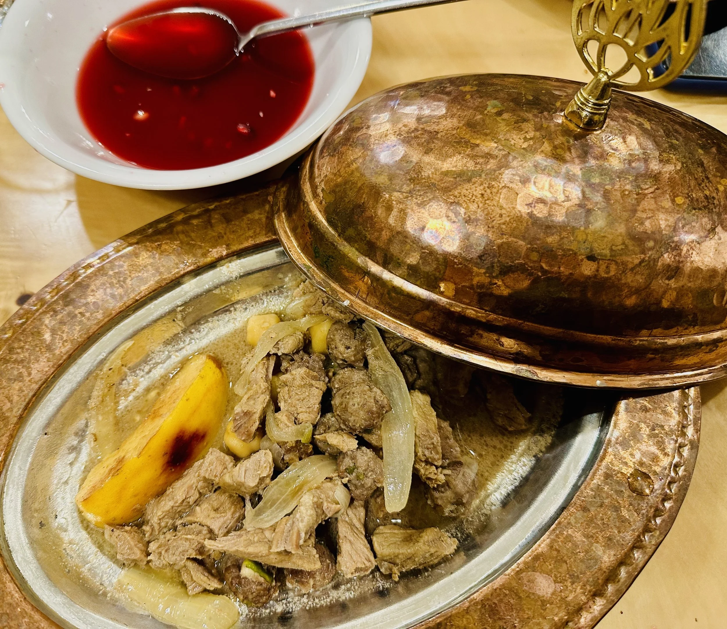 Shepherd's pie served in a copper pan with a lid partially covering the dish, accompanied by a bowl of red jelly or jam. The shepherd's pie contains meat, onions, and a baked potato wedge, with a creamy sauce visible inside.