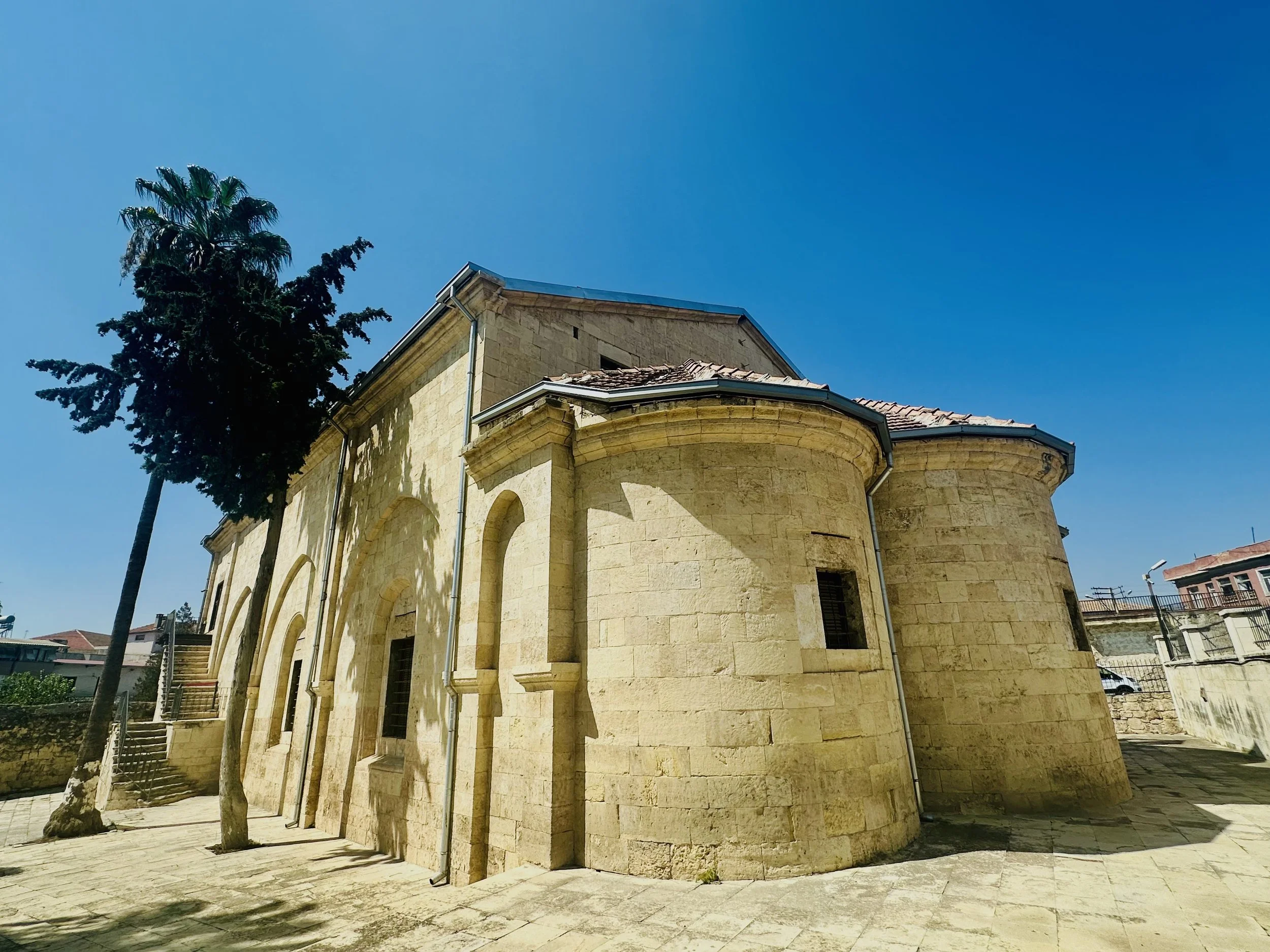 A historic stone church with rounded walls and small windows under a bright blue sky, with palm and other trees nearby.