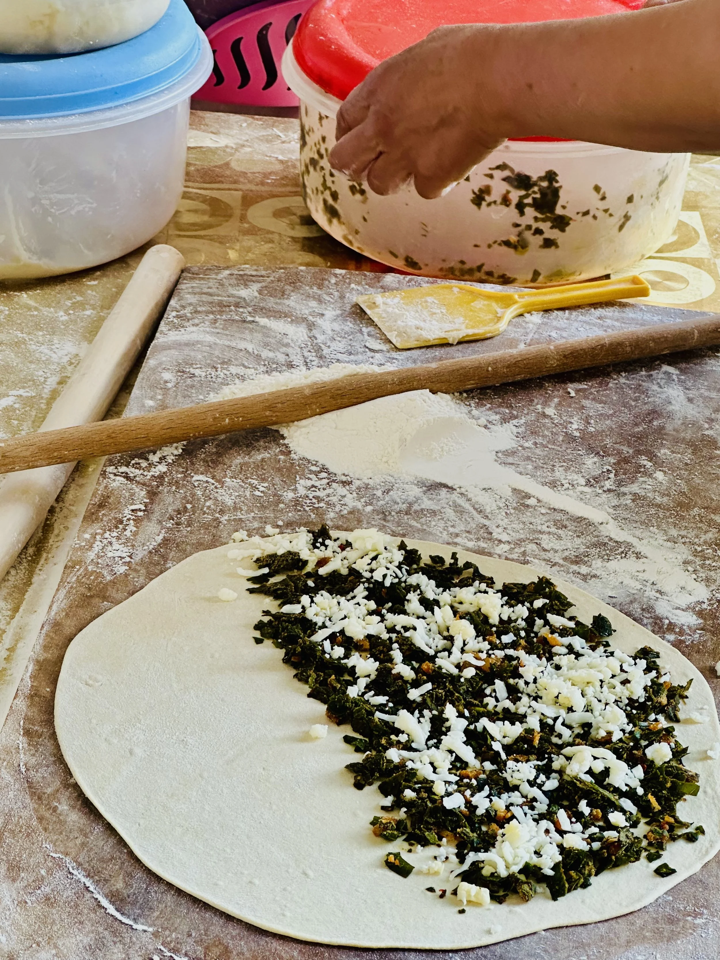 A pizza dough with a spinach and cheese filling on a floured surface, with rolling pins and containers in the background.