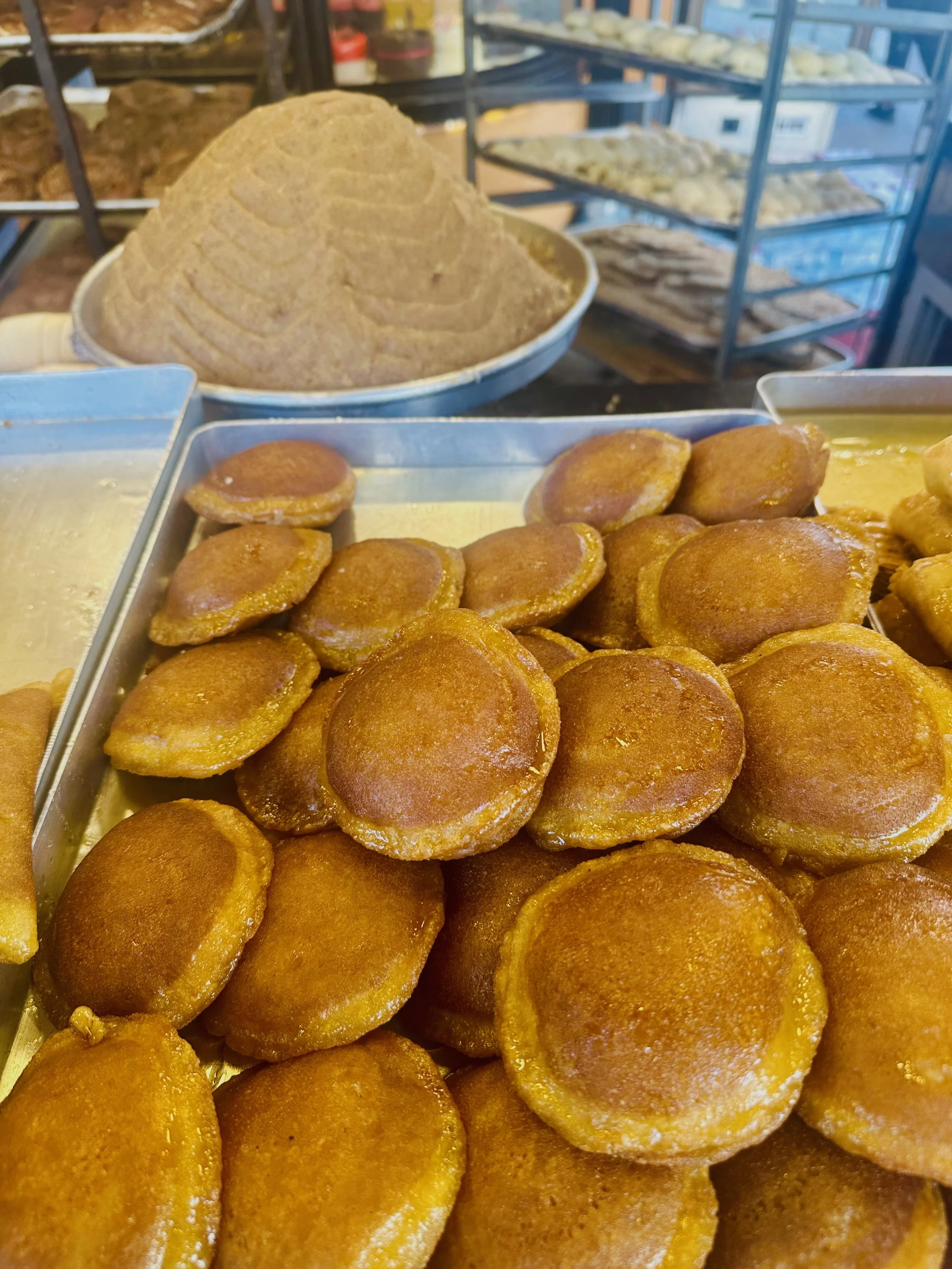 Close-up of fried dough balls in a display tray, with a large mound of what appears to be brown sugar or coarse spice in a bowl behind them, in a bakery or food market setting.