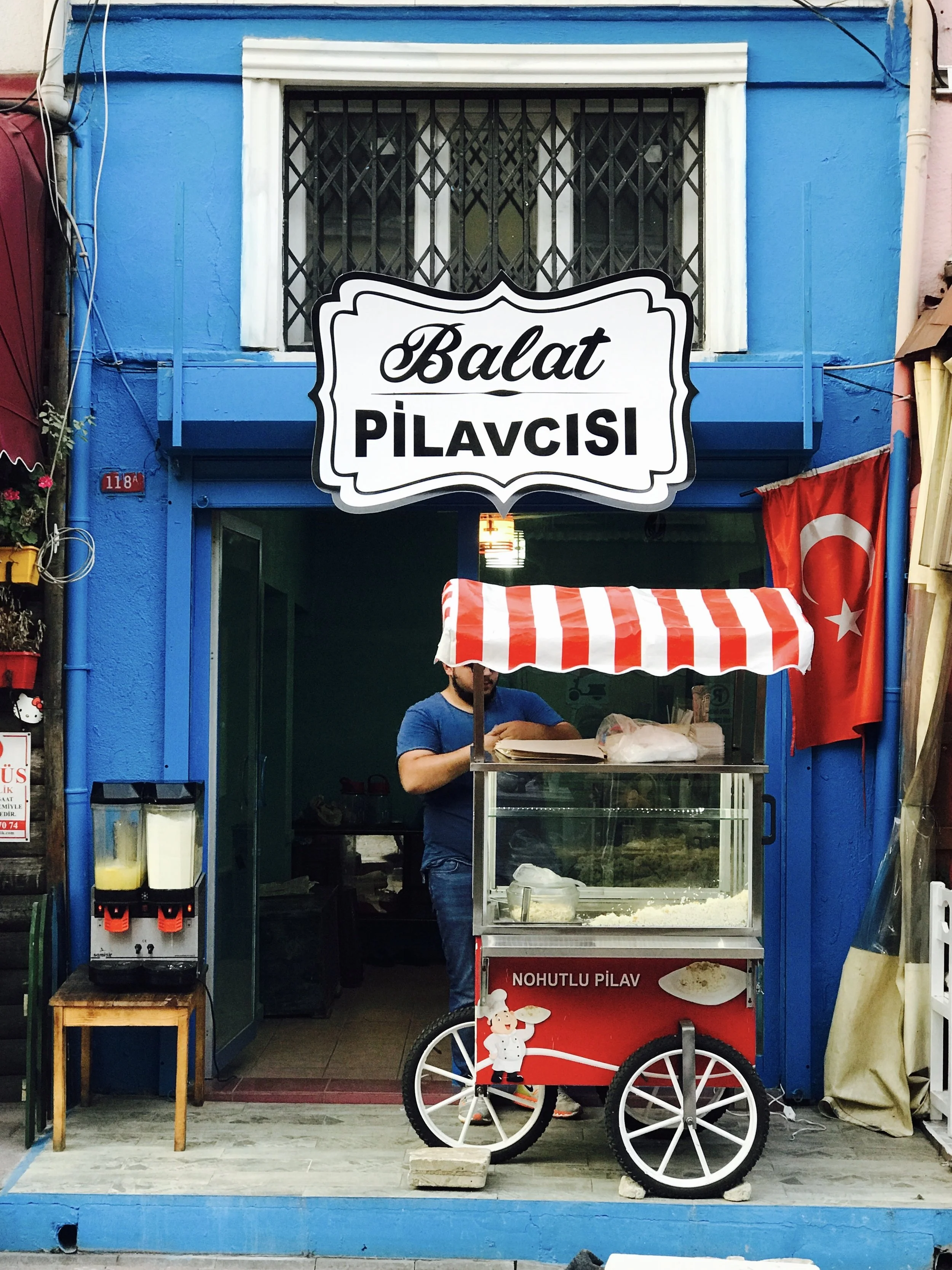 A street food stand selling Balat pilavı, a Turkish rice dish, with a red and white striped canopy, a glass display case, and a person preparing food inside. The stand has a sign with the words "Balat PİLAVCISI" above the entrance, and a Turkish flag on the right side.