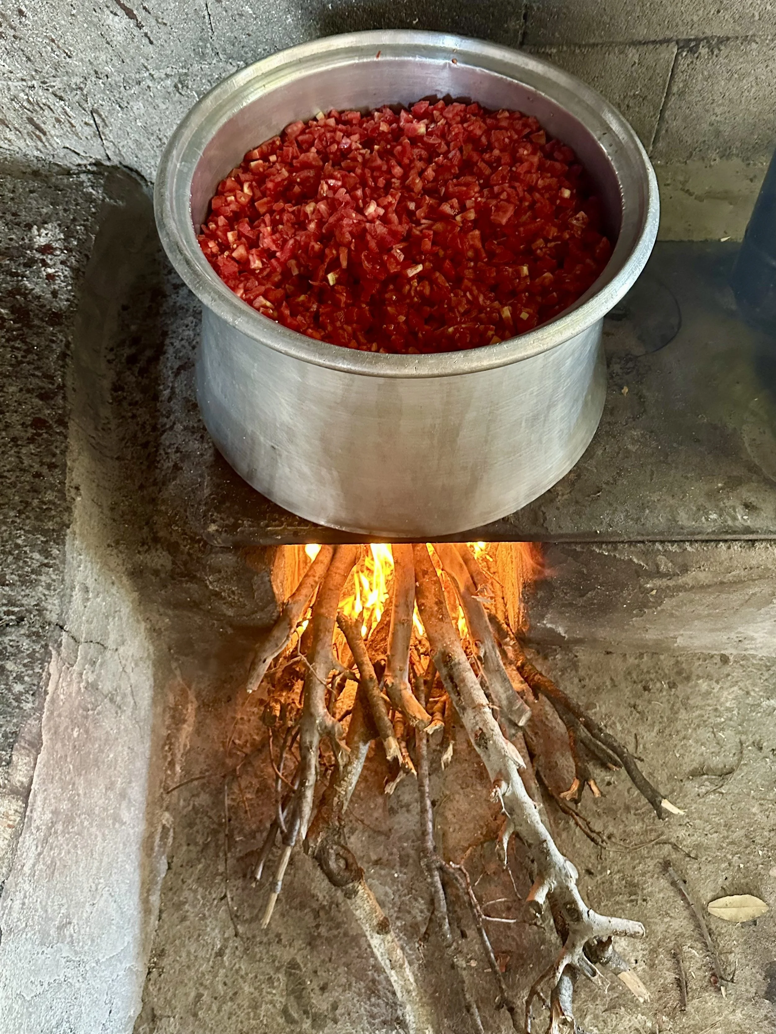 A large metal pot filled with chopped red tomatoes on an outdoor stove with a wood fire made of sticks burning underneath.