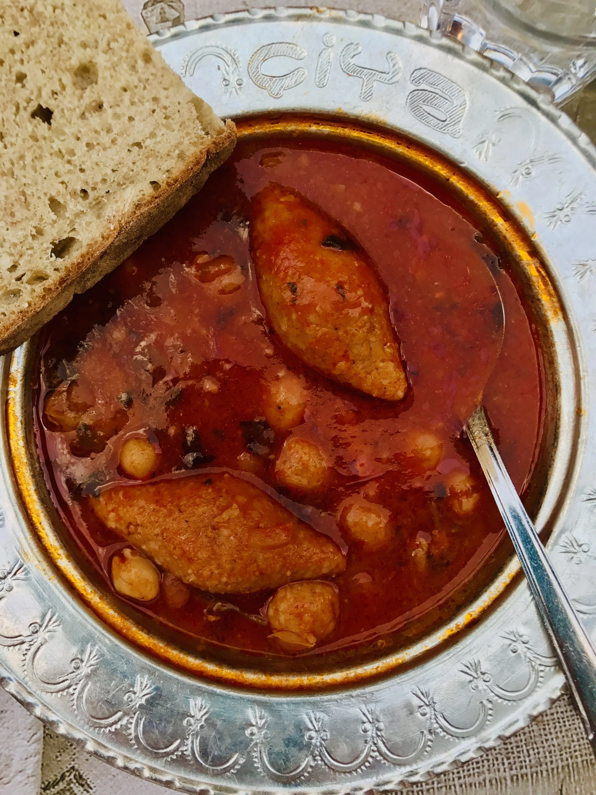 A bowl of tomato-based chicken and chickpea soup with two pieces of bread on the side, served in a silver dish with decorative edges.