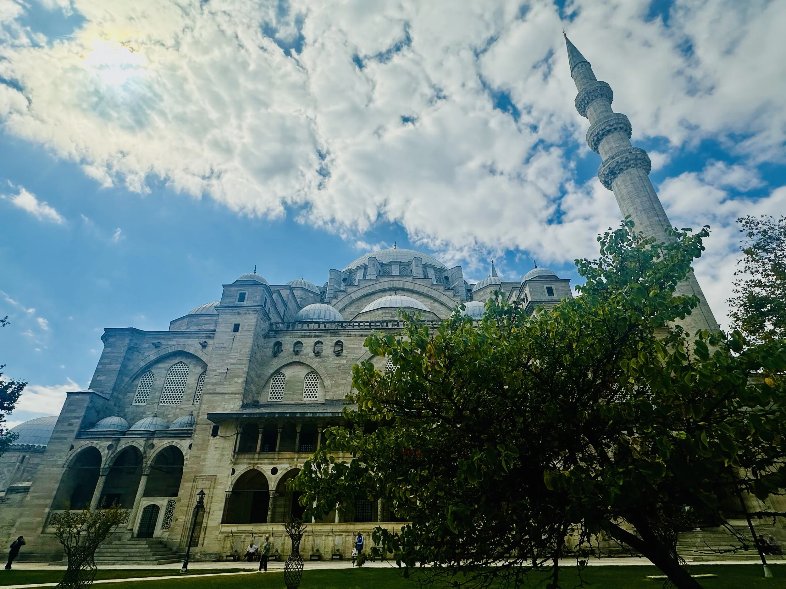 A large mosque with multiple domes and a tall minaret against a partly cloudy blue sky. There are trees and people in the foreground.