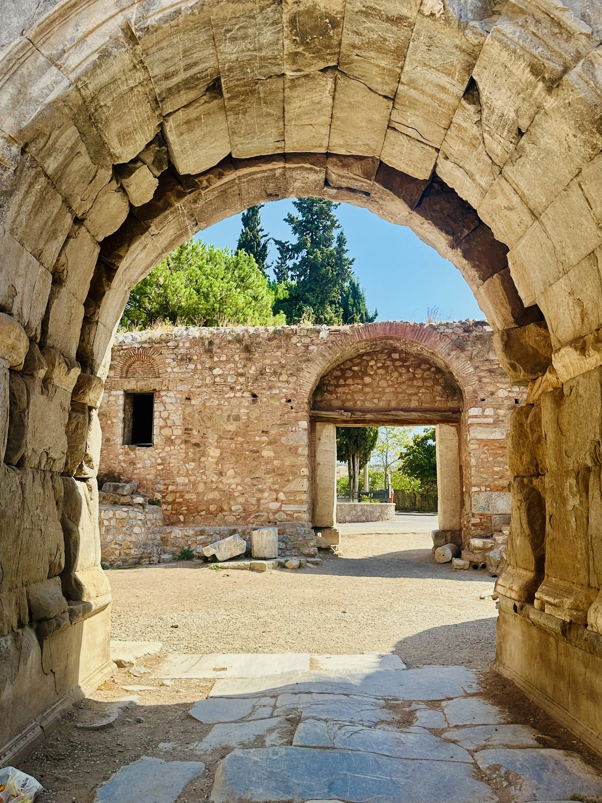 Ancient stone archway leading to a courtyard with a brick wall and trees in the background, and a clear blue sky.