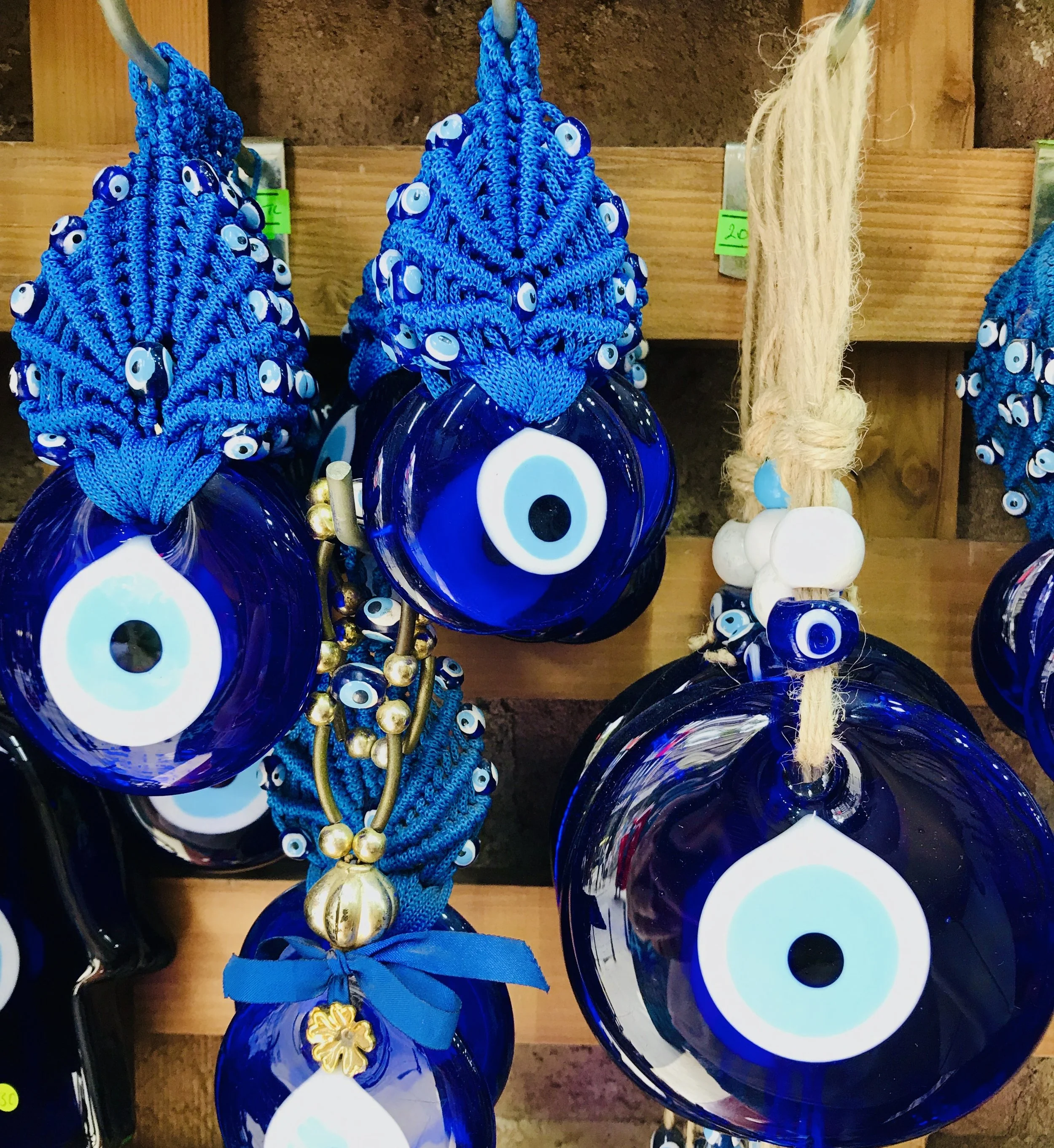 Decorative glass ornaments in dark blue with white and light blue eye motifs, hanging from blue braided cords and beige rope, adorned with gold and white beads.