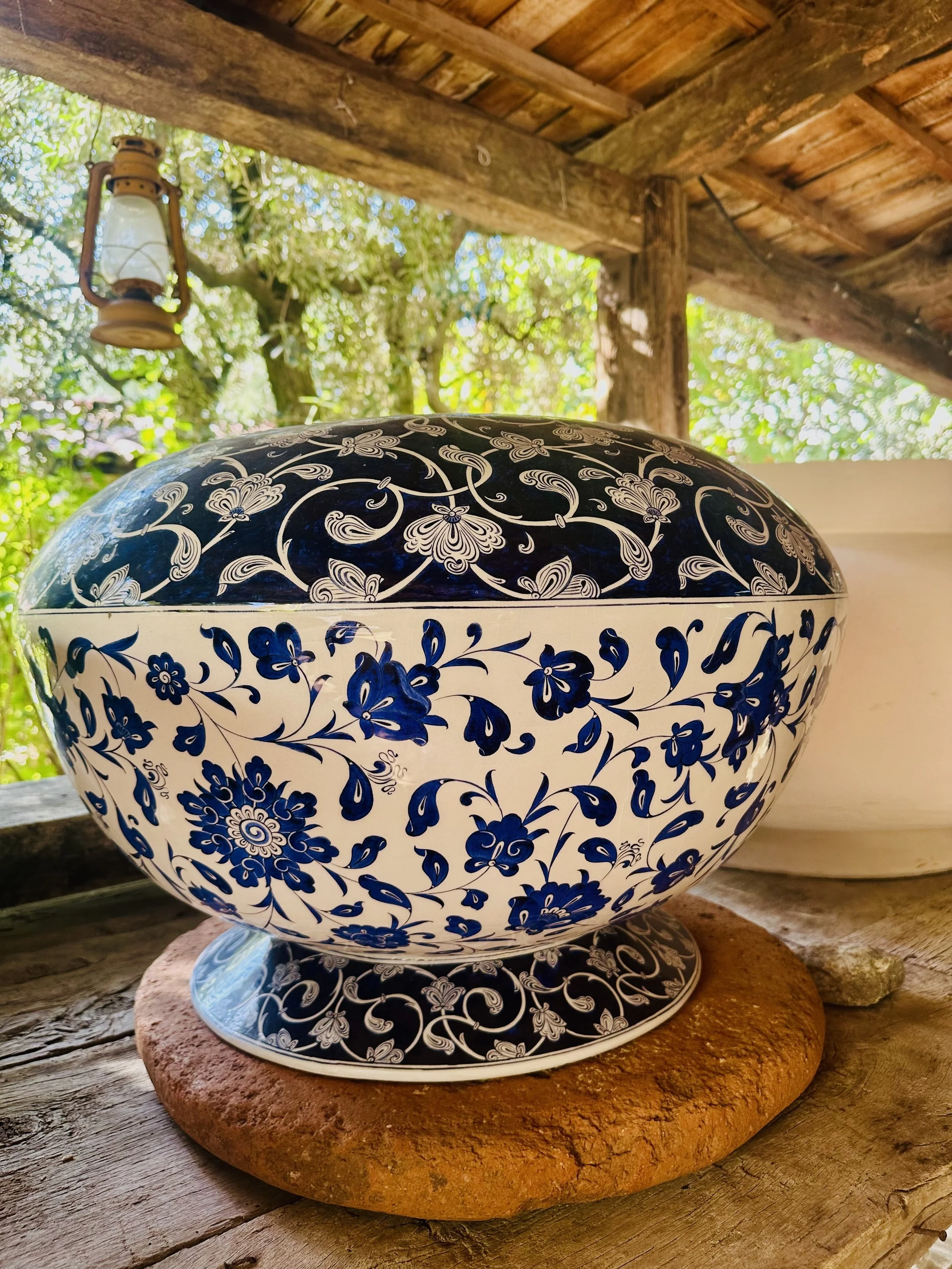 Decorative ceramic bowl with blue floral patterns on a white background, resting on a piece of red brick, in an outdoor setting with a wooden roof and lantern in the background.