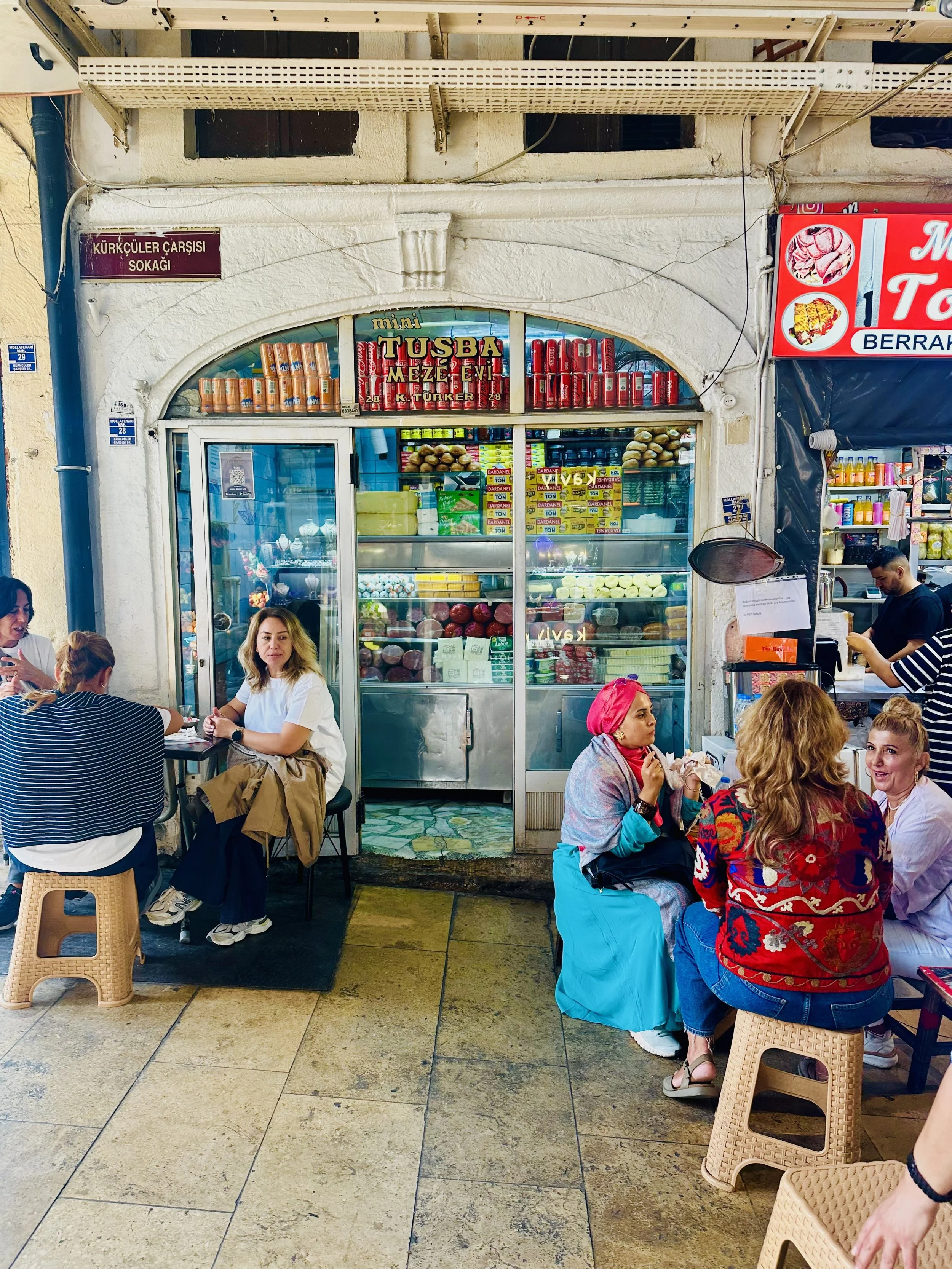 Outdoor cafe with people sitting at tables in front of a small shop selling food and snacks. The shop window displays fruits and packaged items. Signs with Turkish text are visible, indicating it is in Turkey.