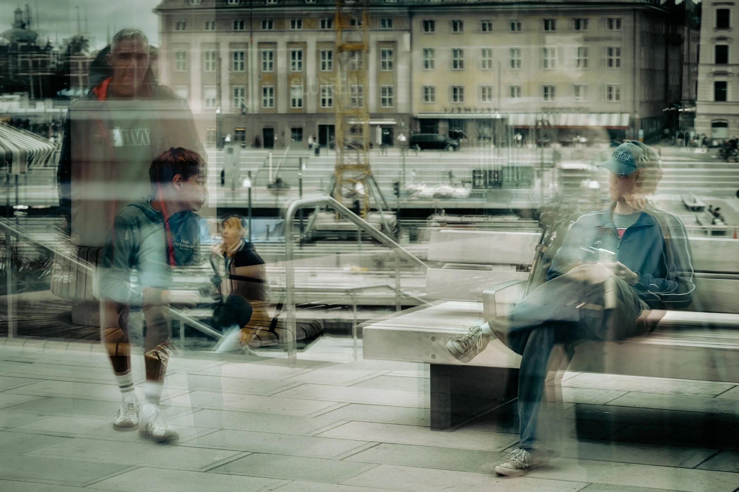 In einem Wartebereich sitzende Menschen spiegeln sich in großen Fenstern wider, im Hintergrund sind Stadtgebäude und Baukräne zu sehen.