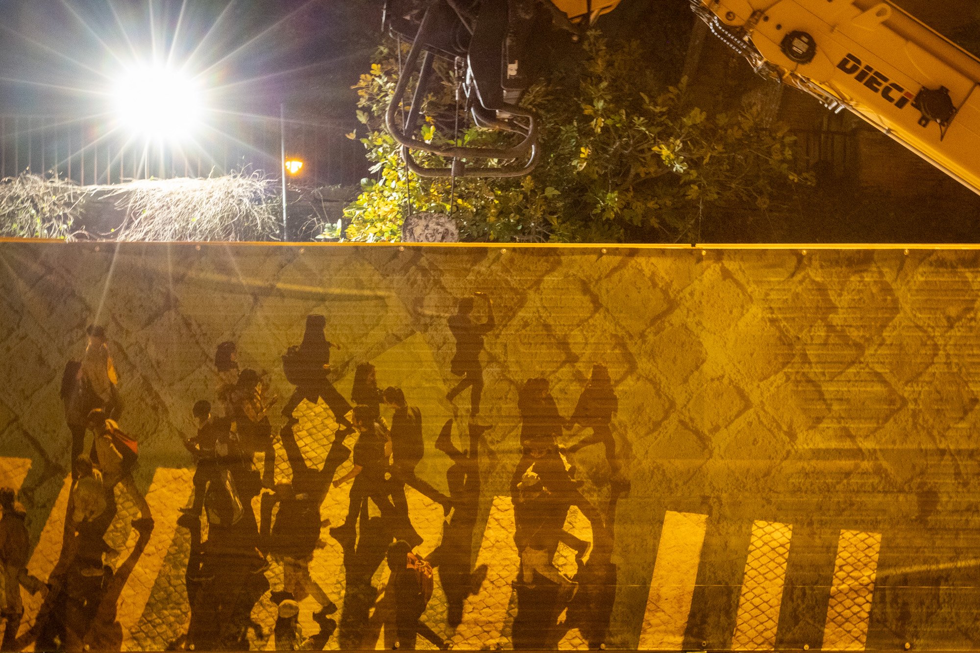 Nachtfoto eines beleuchteten Parks mit den Schatten von Menschen, die über eine Steinmauer laufen, und einem Kran im Hintergrund.